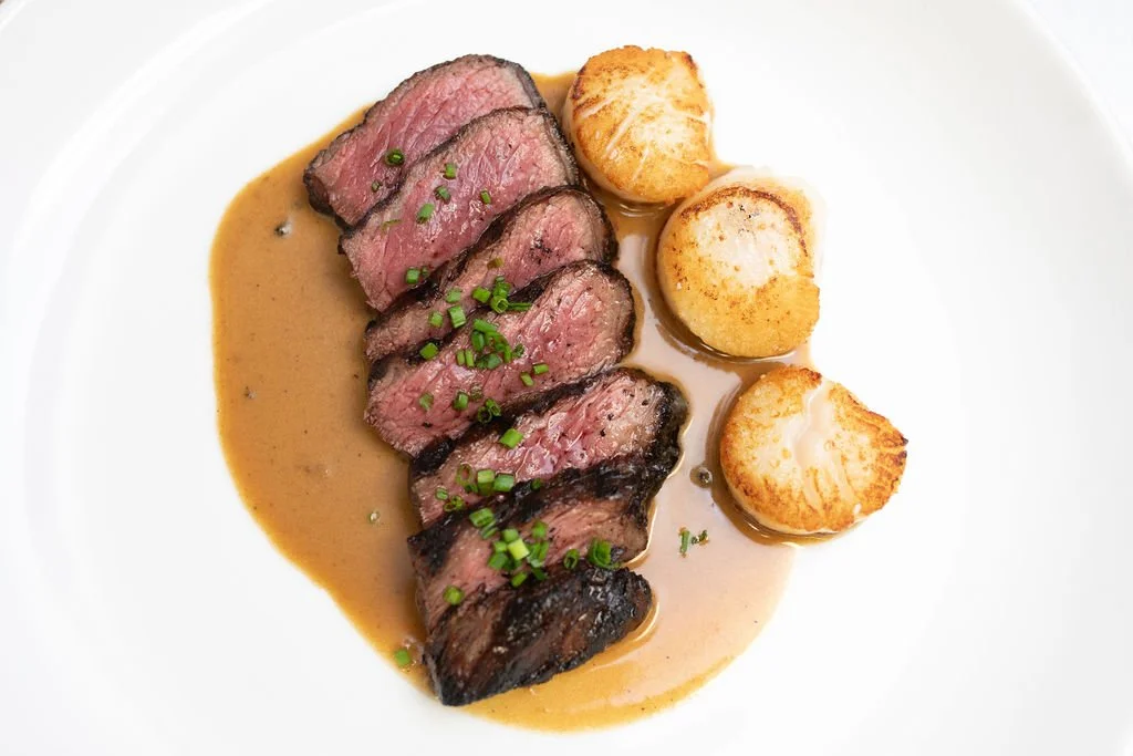 Sliced cooked beef steak topped with chopped chives, served with three seared scallops on a white plate with sauce