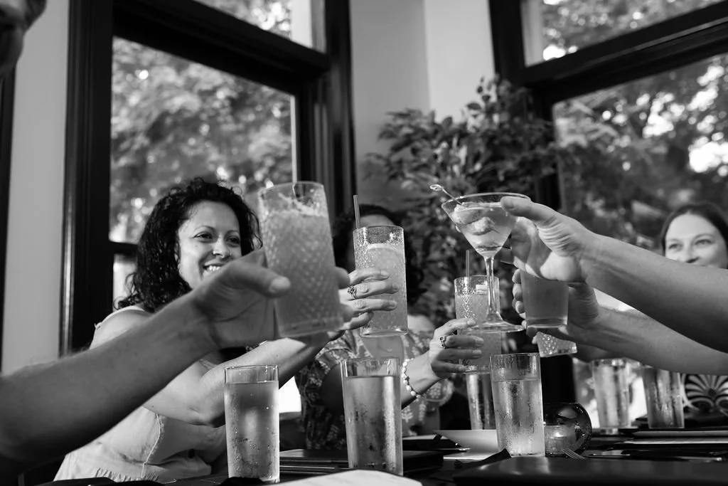 People raising glasses in a toast at a celebration at Table 1888, sitting at a table with drinks, in a bright room with large windows and trees outside.