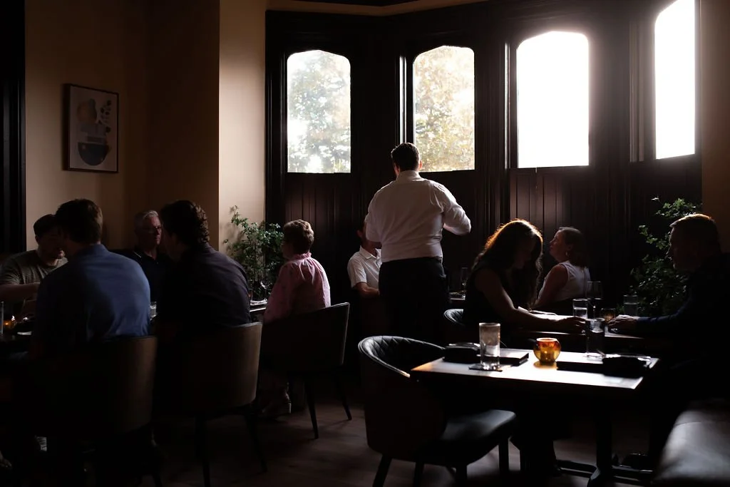 People dining in a dimly lit restaurant with large windows letting in natural light, and a waiter serving a table.