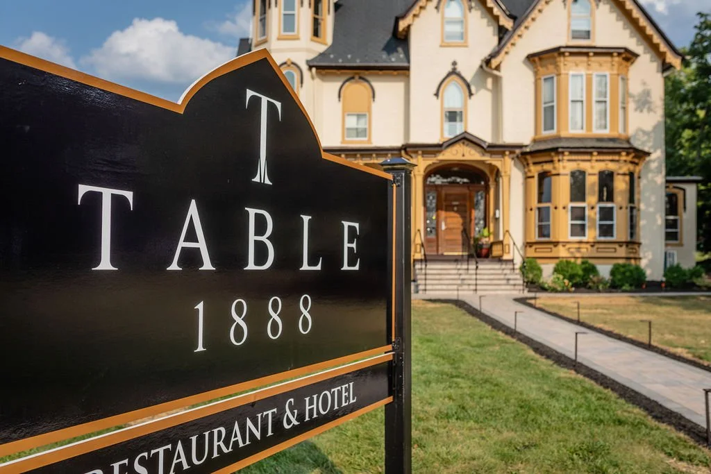 A large black sign with white text reading "T TABLE 1888" and "RESTAURANT & HOTEL" in front of a Victorian-style building with yellow and brown trim, set on a green lawn with a stone pathway leading up to the entrance.