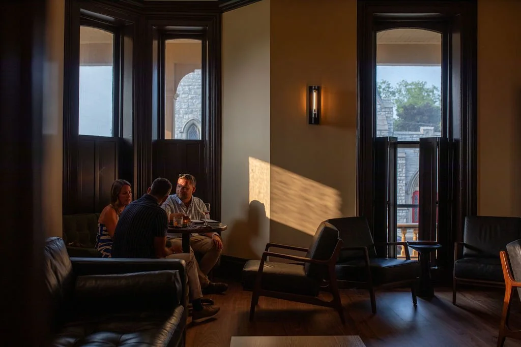 Three people sitting around a small table near large windows with daylight streaming in, inside a dimly lit room with dark wood furniture.
