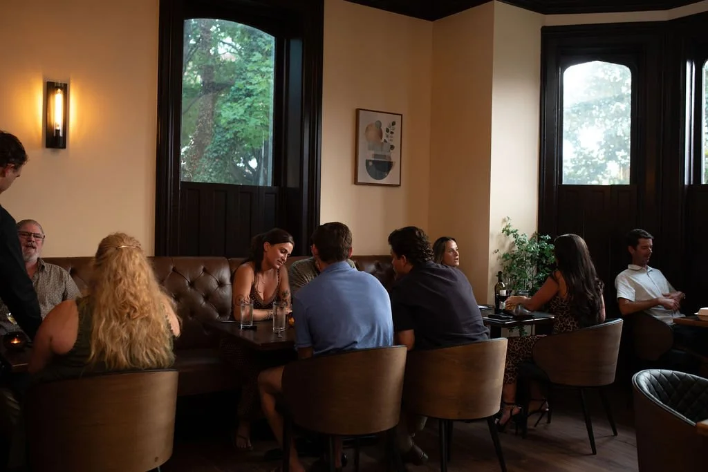 People dining in a cozy restaurant with large windows, a leather bench, and artwork on the wall.