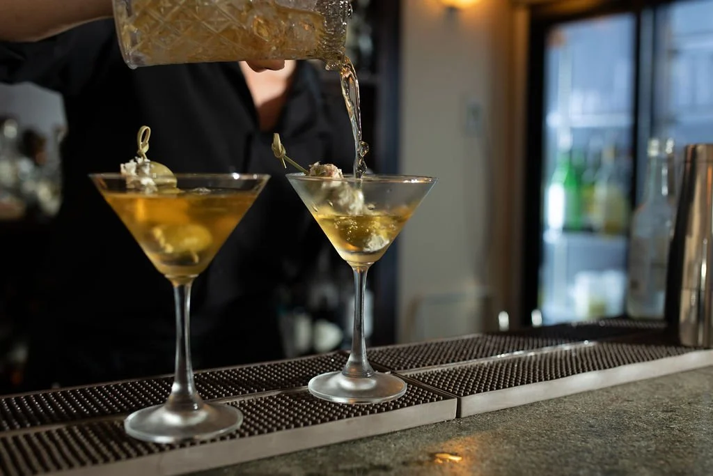 A bartender pours a drink into two martini glasses with garnishes at a bar.