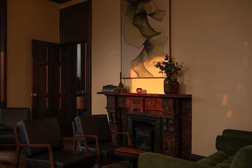 A dimly lit living room with a dark wooden fireplace mantle, green and black armchairs, a painting above the fireplace, and a potted plant with flowers on top of the mantle.