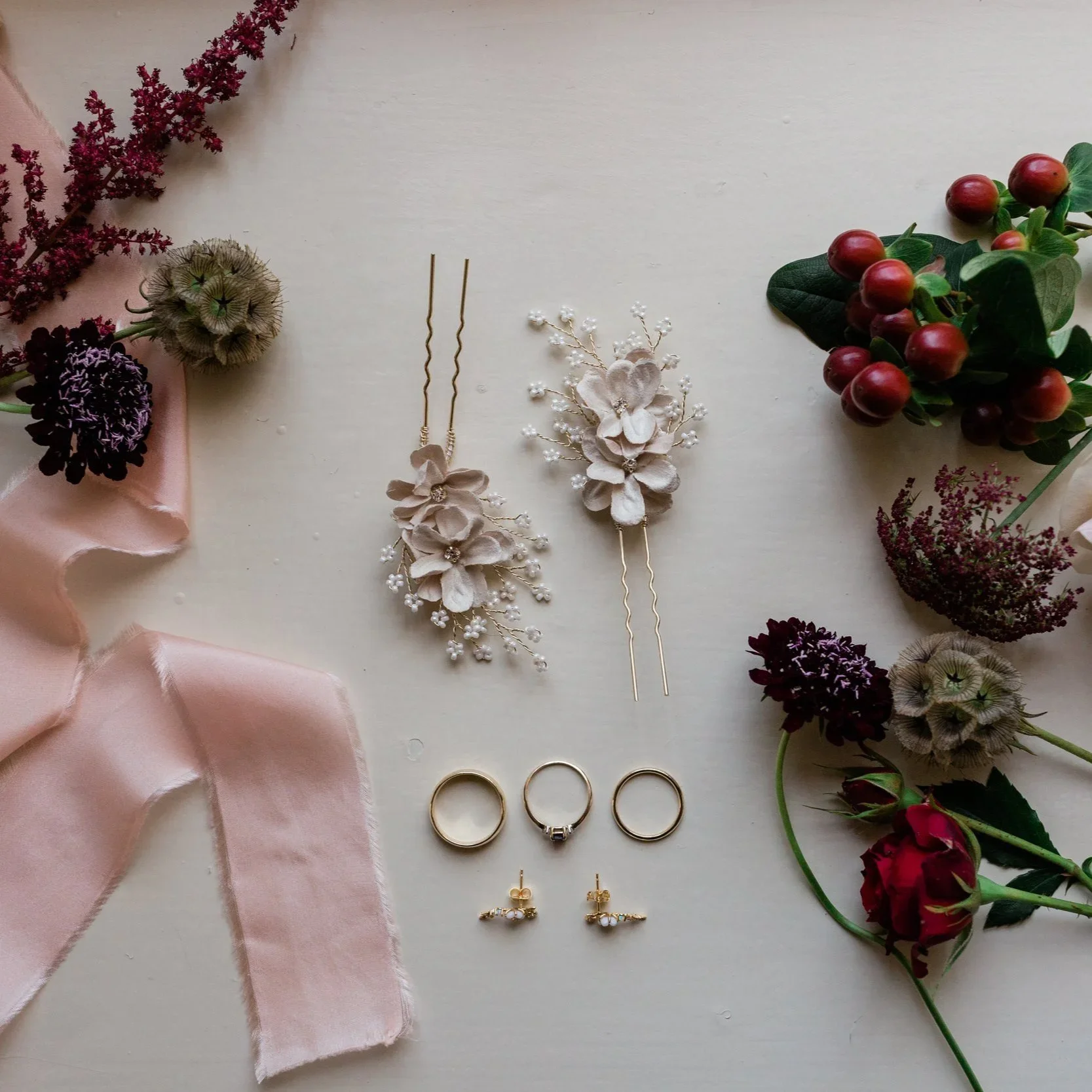Flatlay of wedding jewelry including a hairpins, earrings, and wedding rings, surrounded by dried flowers and a pink ribbon on a white surface.