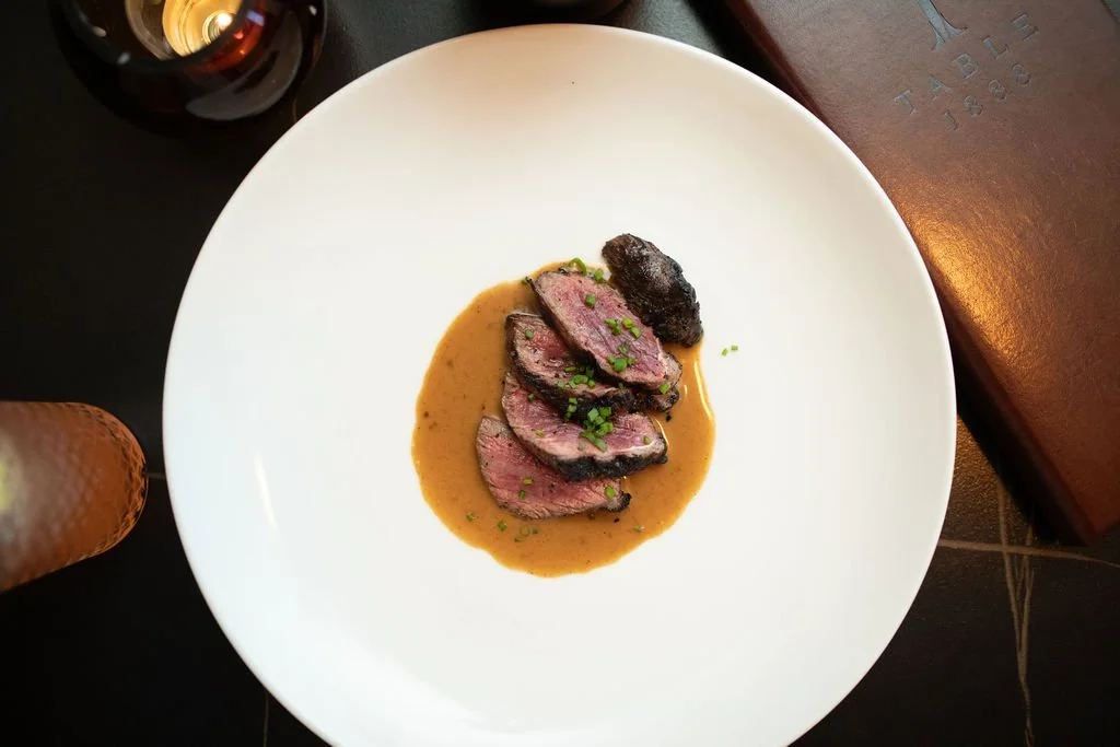 Slices of cooked beef steak with a dark crust, served with brown gravy and garnished with chopped green herbs, on a large white plate.
