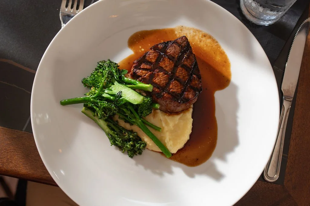 Grilled steak with grill marks served with green broccoli and mashed potatoes, topped with brown gravy, on a white plate.