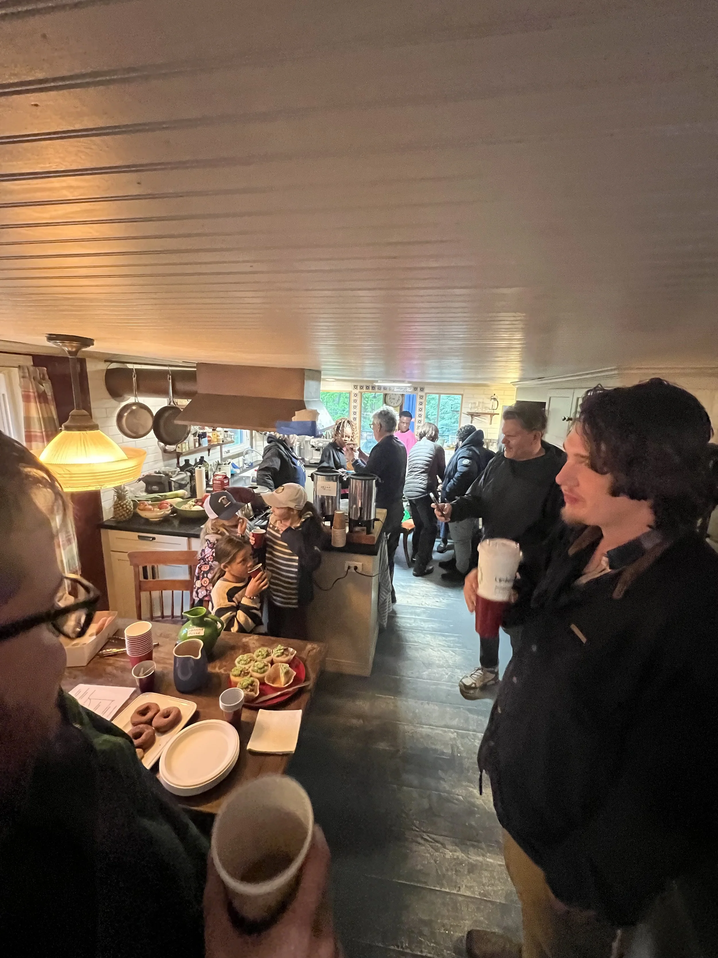 People gathered in a cozy kitchen for a casual social gathering, with snacks and drinks on the counter and everyone holding cups.