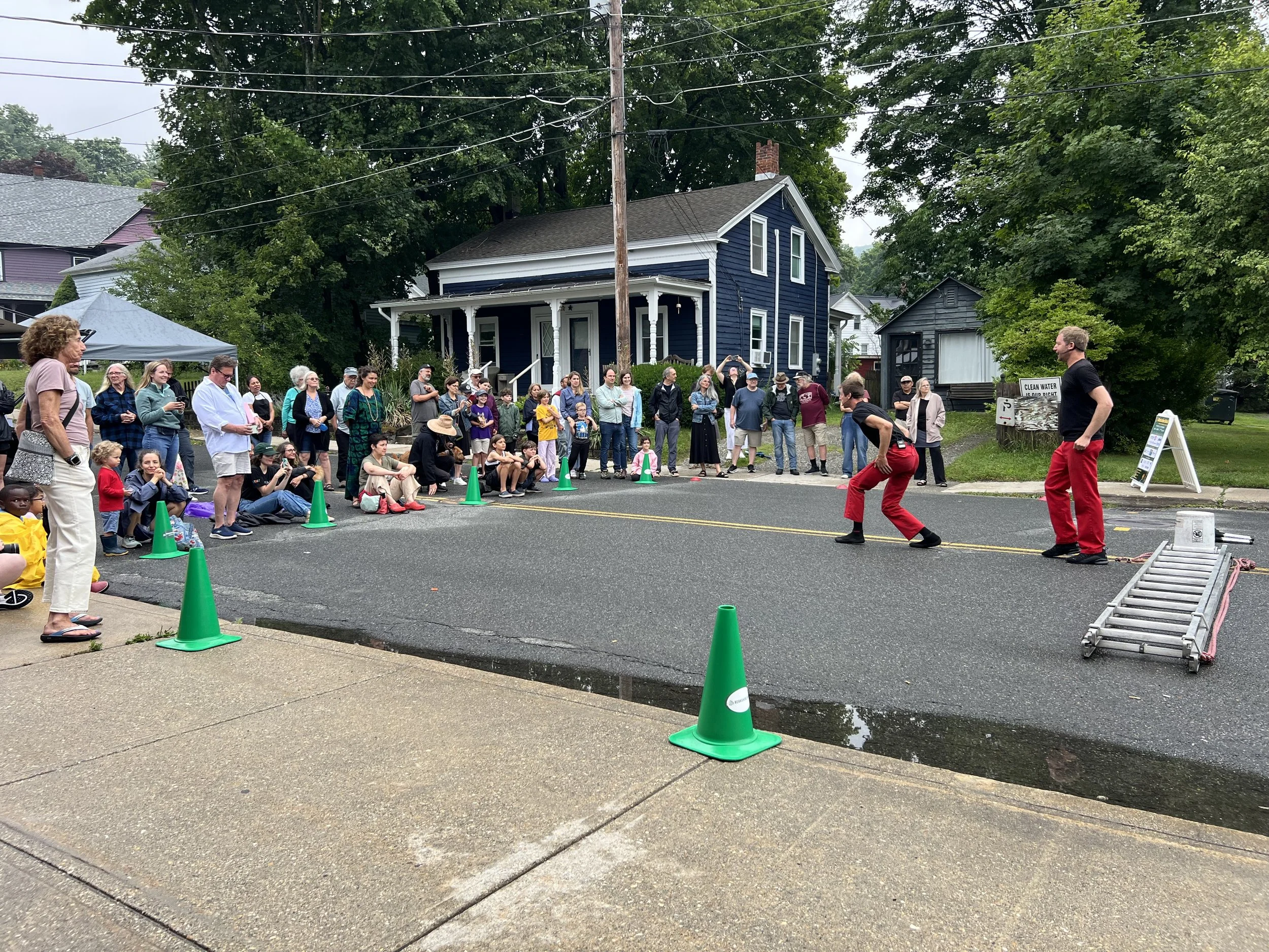 A crowd of people gathered on a street corner or sidewalk, watching two performers dressed in red pants and black shirts in a street performance. There are green cones in the street marking boundaries, and a ladder lying on the sidewalk. In the background, there are houses with trees and power lines.