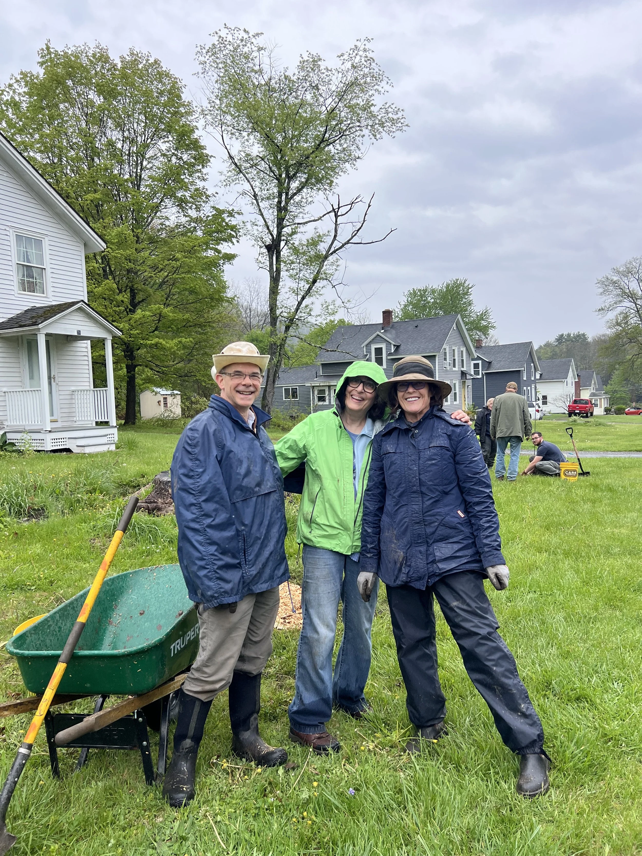 Three people standing outdoors in a grassy area, smiling at the camera. They are dressed in rain jackets, waterproof boots, and hats. Behind them, there are several houses and other individuals engaged in garden or yard work, with tools and equipment visible, on a cloudy day.