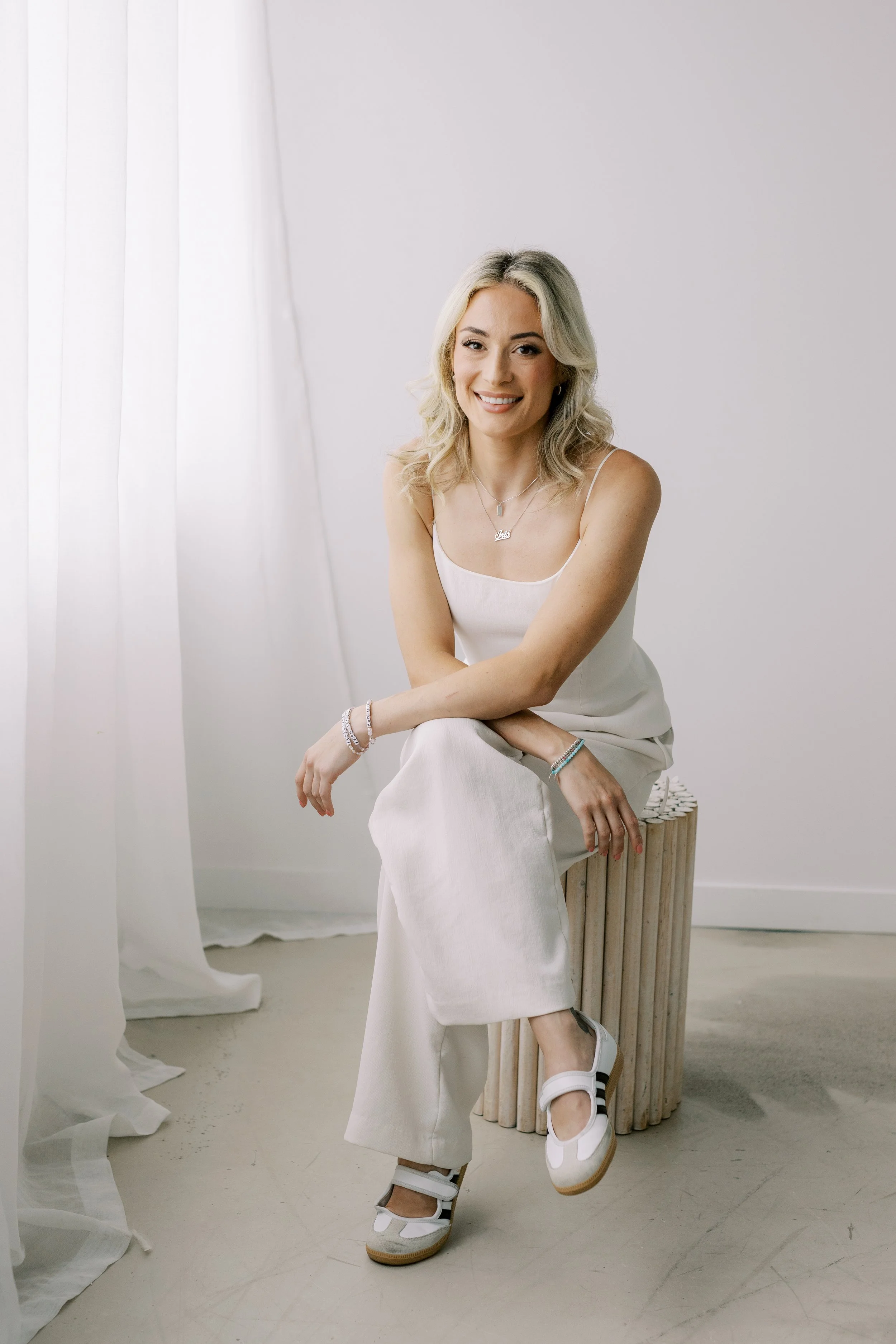 Smiling woman with blonde hair sitting on a wooden stool in a minimalist white room, wearing a white dress and sneakers, with a white curtain and plain wall background.