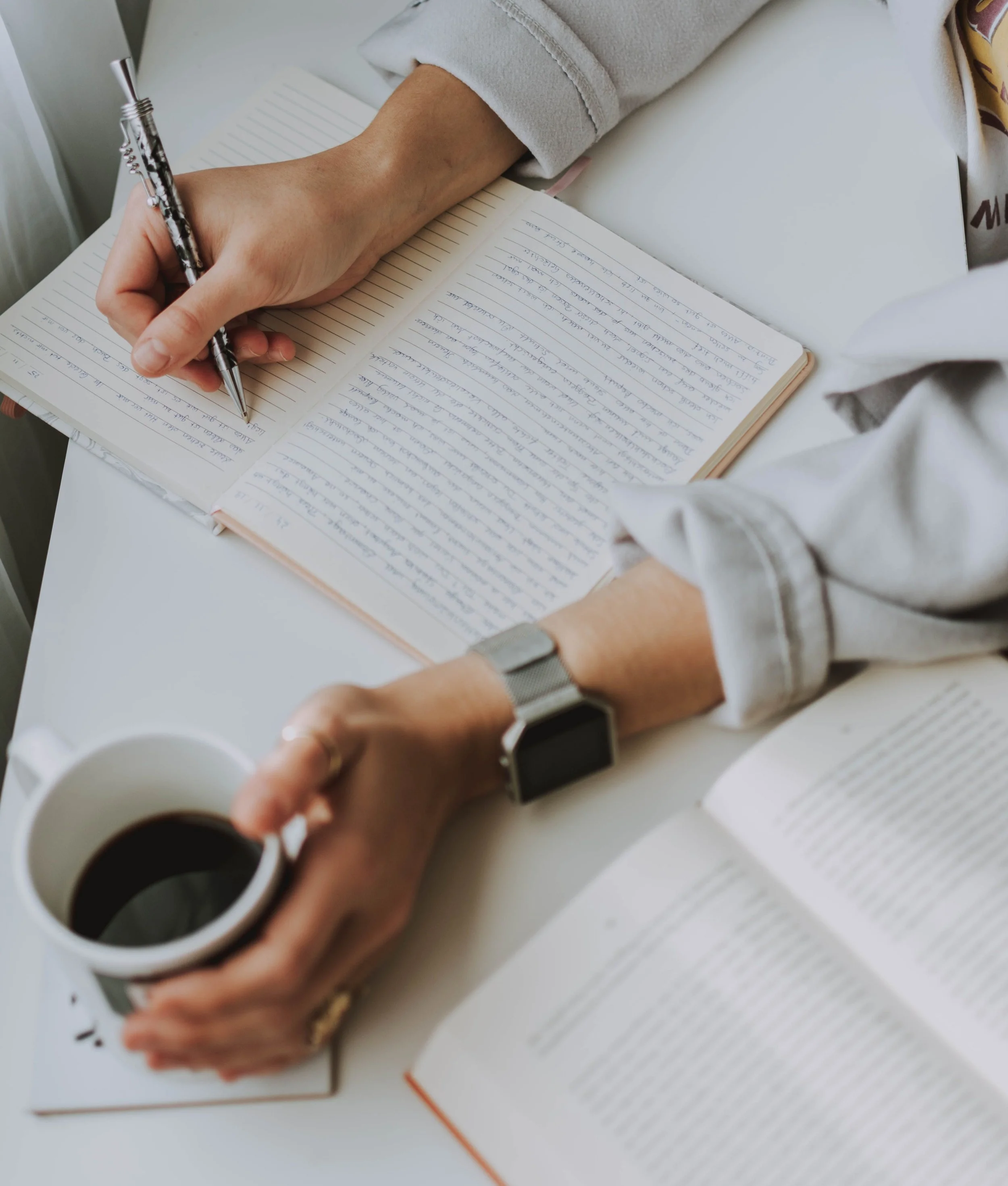 Person writing in a notebook with a patterned pen, wearing a watch, next to a cup of coffee on a white table.