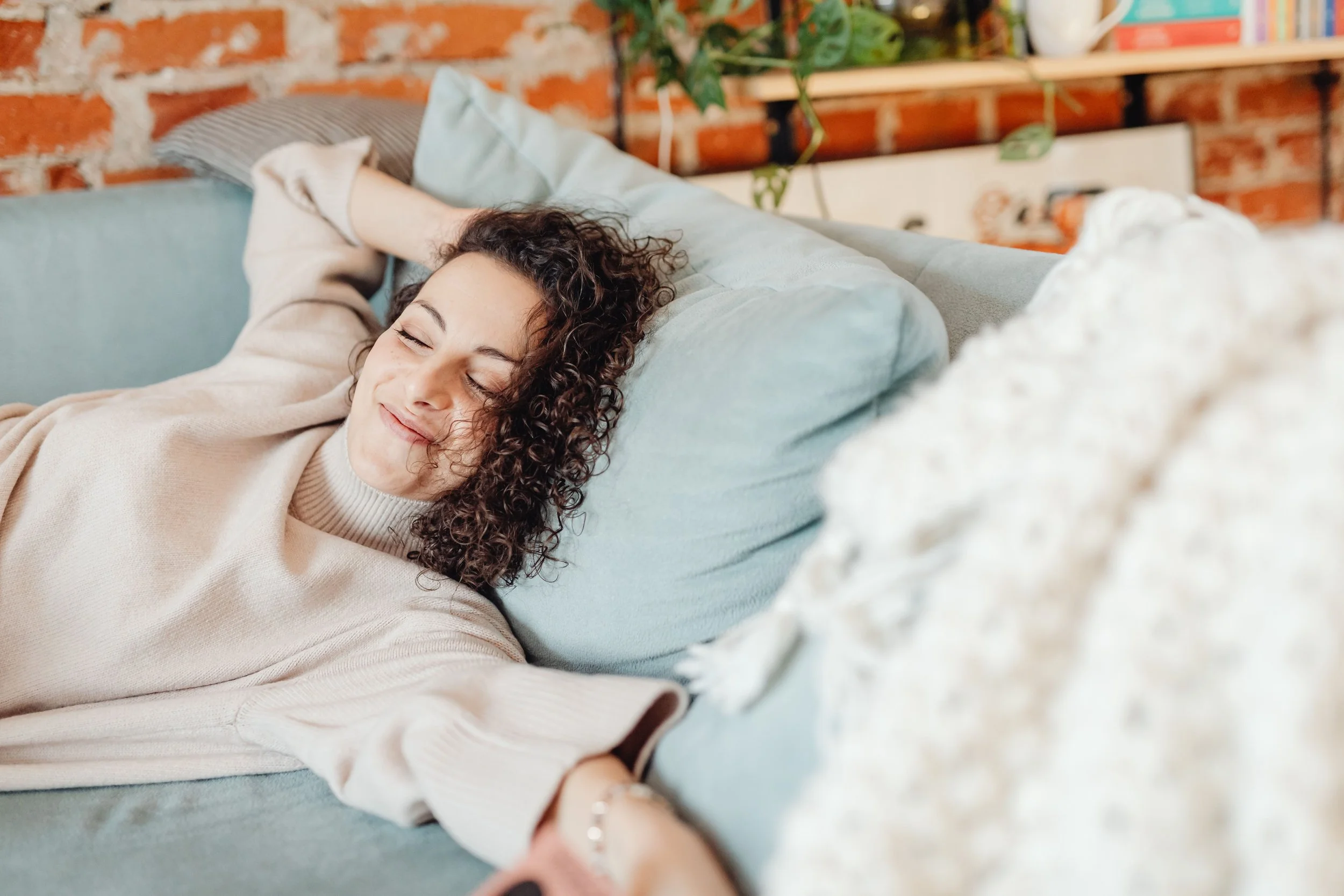 A woman with curly hair lying on a light green sofa, smiling with eyes closed, in a cozy room with brick walls and wooden shelves in the background.