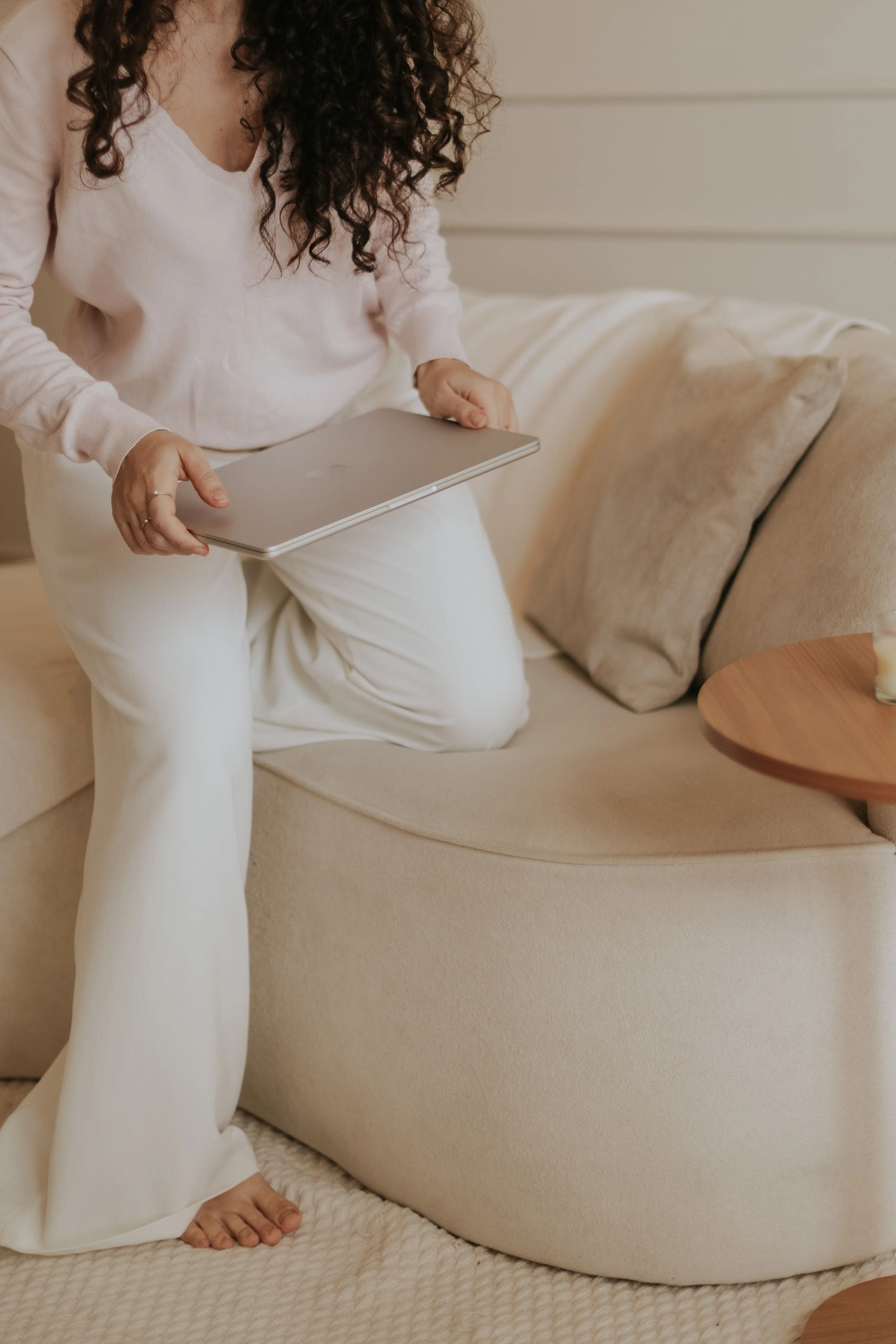 A woman with curly hair is kneeling on a cream-colored rug, holding a laptop, in a cozy living room with beige furniture and pillows.