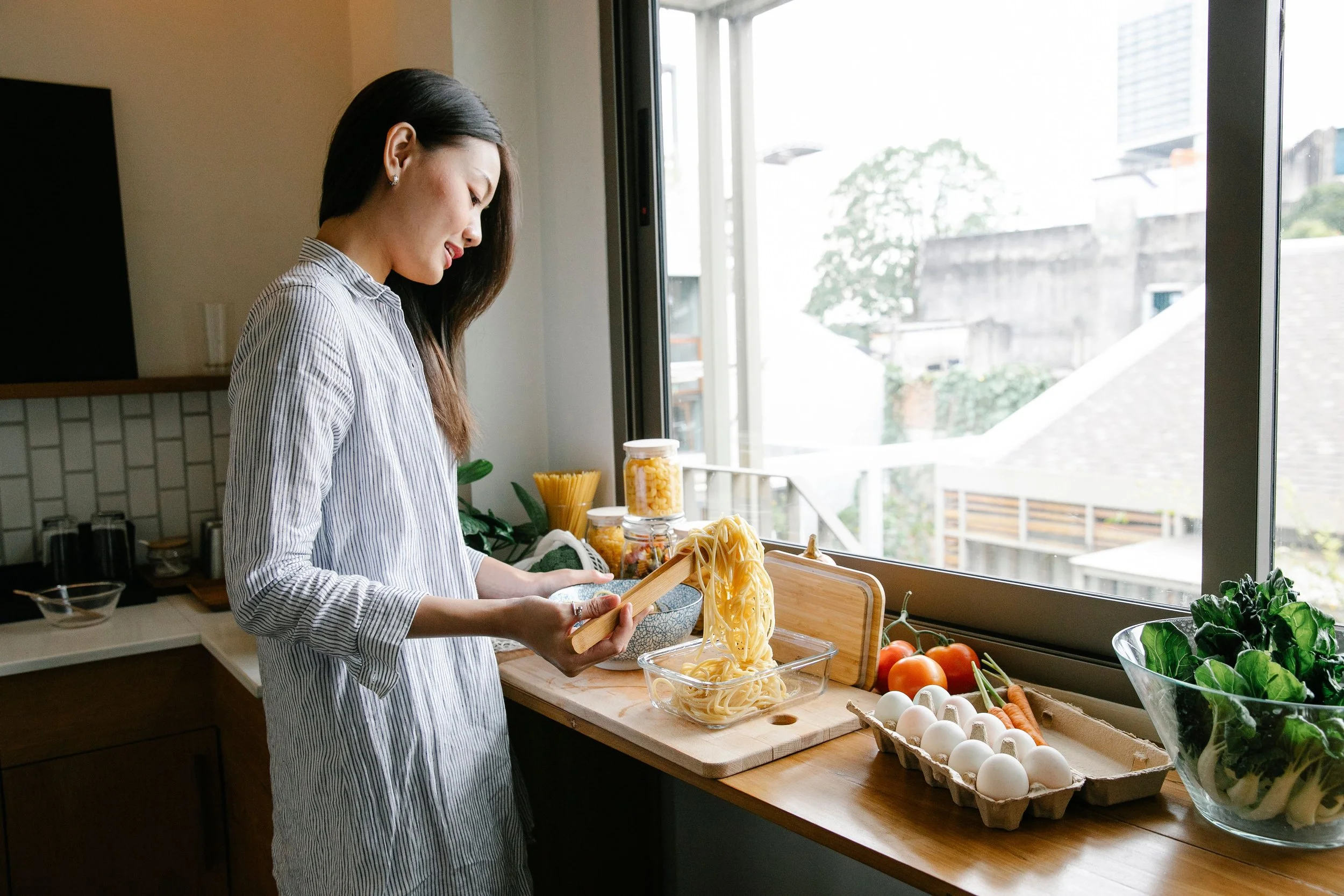 Woman in striped shirt preparing pasta in a bright kitchen near a large window, with fresh vegetables and eggs on the counter.