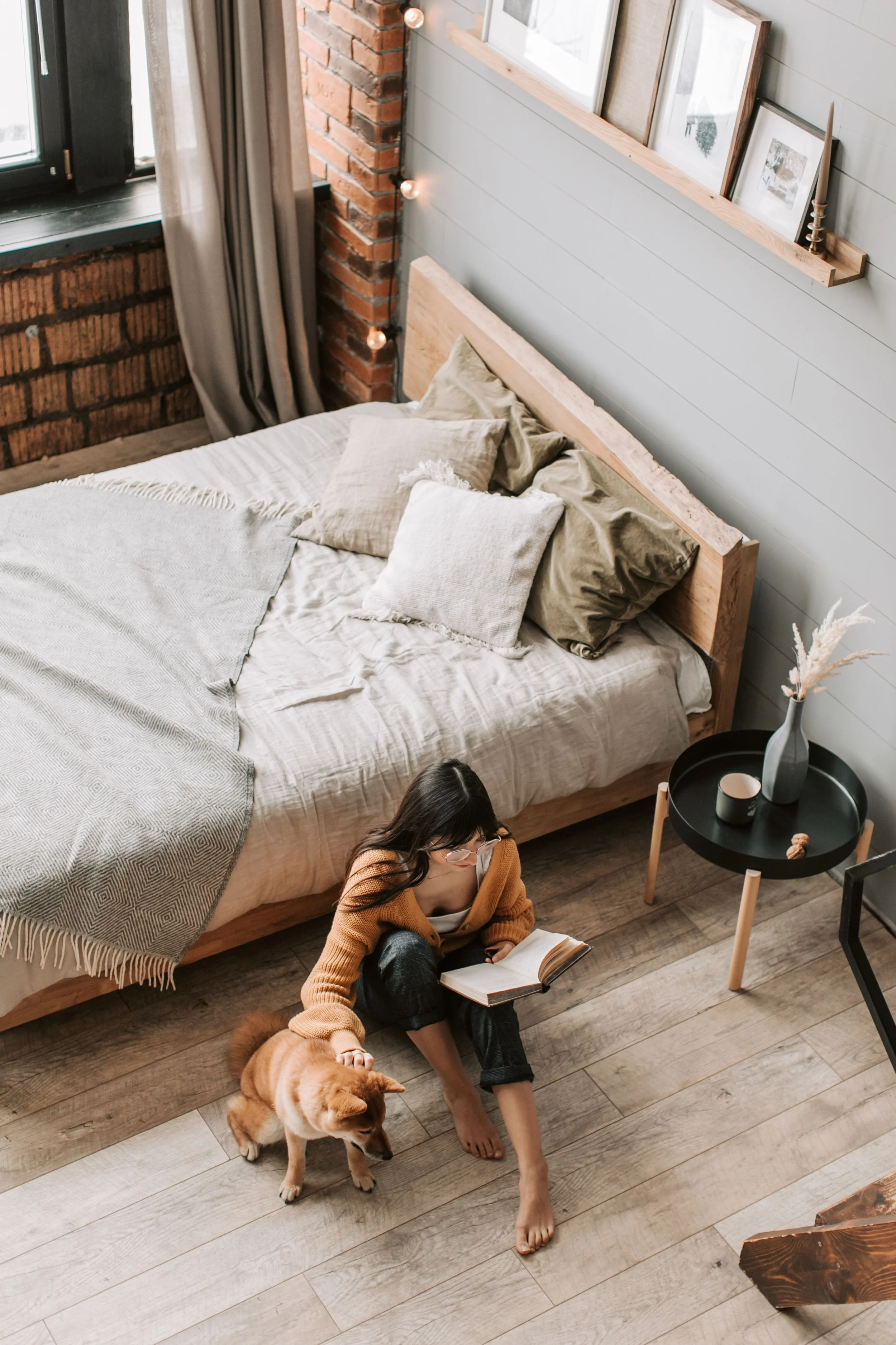 A woman with glasses and dark hair, wearing a tan cardigan and black pants, sits on the wooden floor reading a book. She is barefoot and petting a small, fluffy dog that is standing beside her. The scene takes place in a bedroom with a bed that has beige pillows and a blanket, a black circular side table with a white vase holding dried pampas grass, and a window with gray curtains. On the wall above the bed, there is a wooden shelf with framed pictures and a candlestick. Decorative string lights are hanging along the brick wall beside the window.