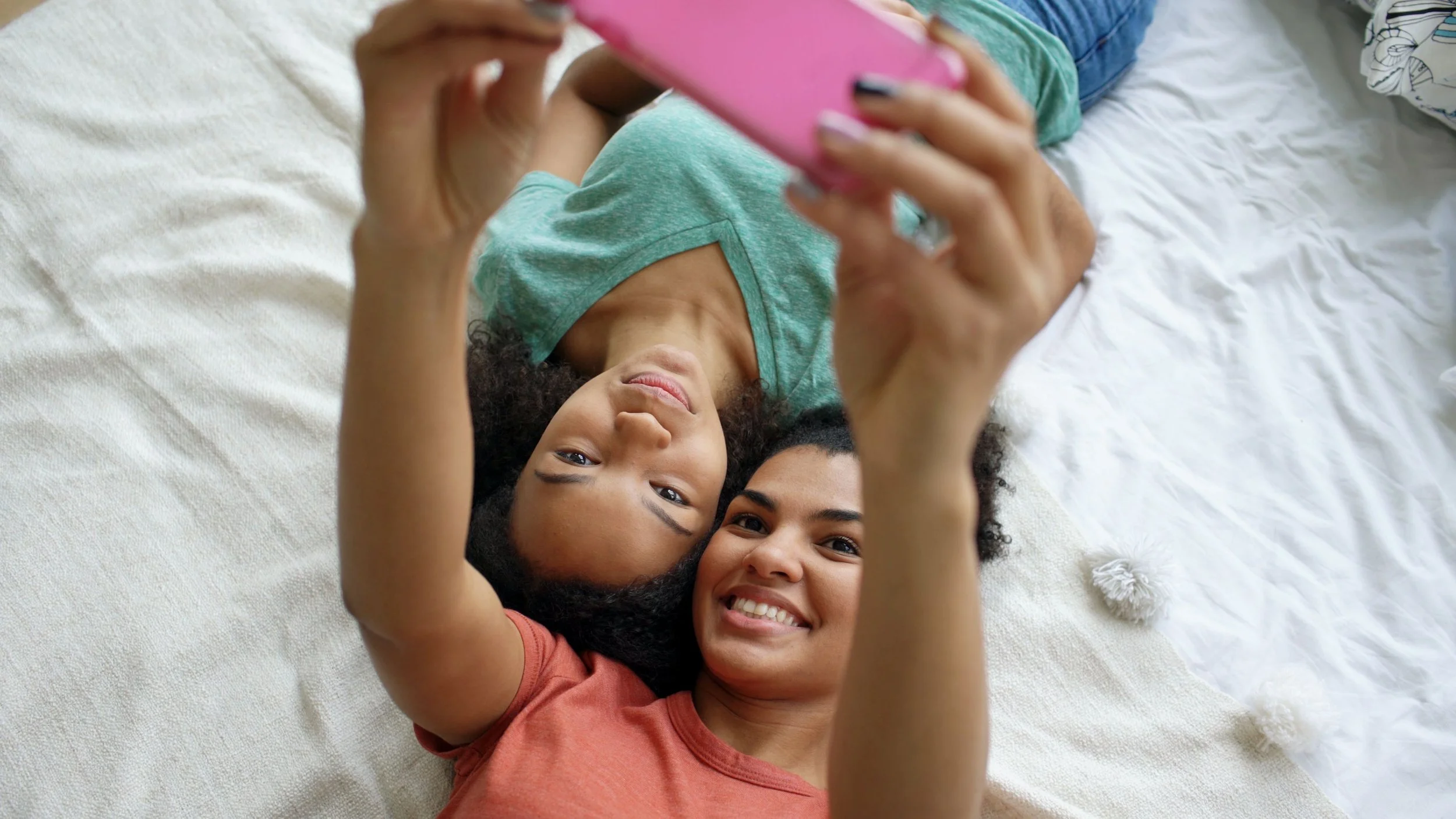Two young women with curly hair lying on a bed, taking a selfie with a pink phone, smiling, and looking at the camera.