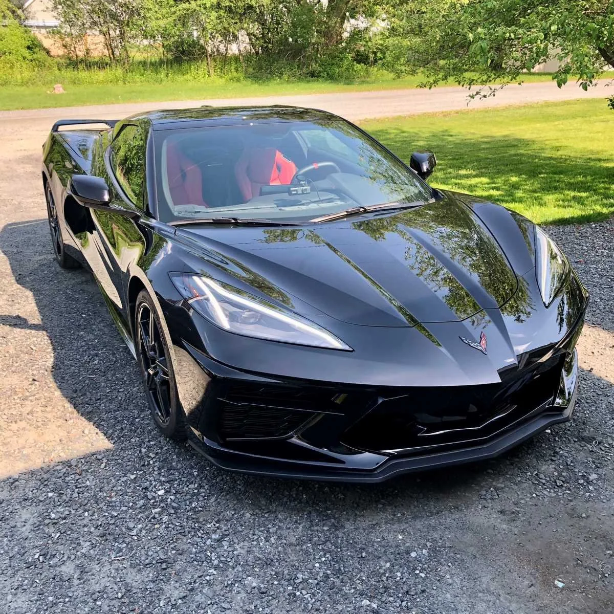 A black sports car parked on a gravel surface, with reflections of trees on its hood and windshield, and red interior seats visible through the windshield.