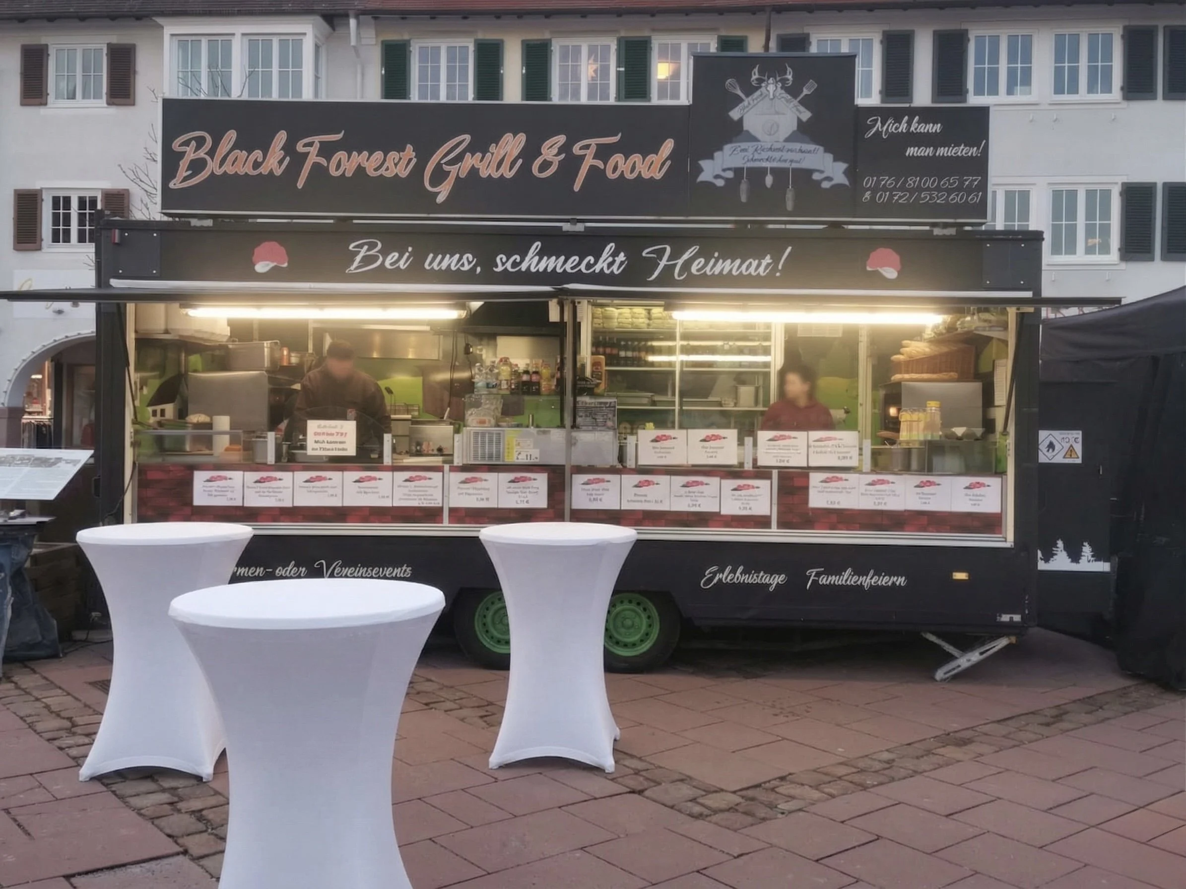 Ein Verkaufswagen mit einem schwarzen Schild darüber, auf dem steht 'Black Forest Grill & Food' und darunter eine Botschaft. Der Wagen hat eine offene Seite mit Speisekarten. Es sind zwei Personen sichtbar, die im Wagen arbeiten. Es gibt auch weiße Stehtische vor dem Wagen.