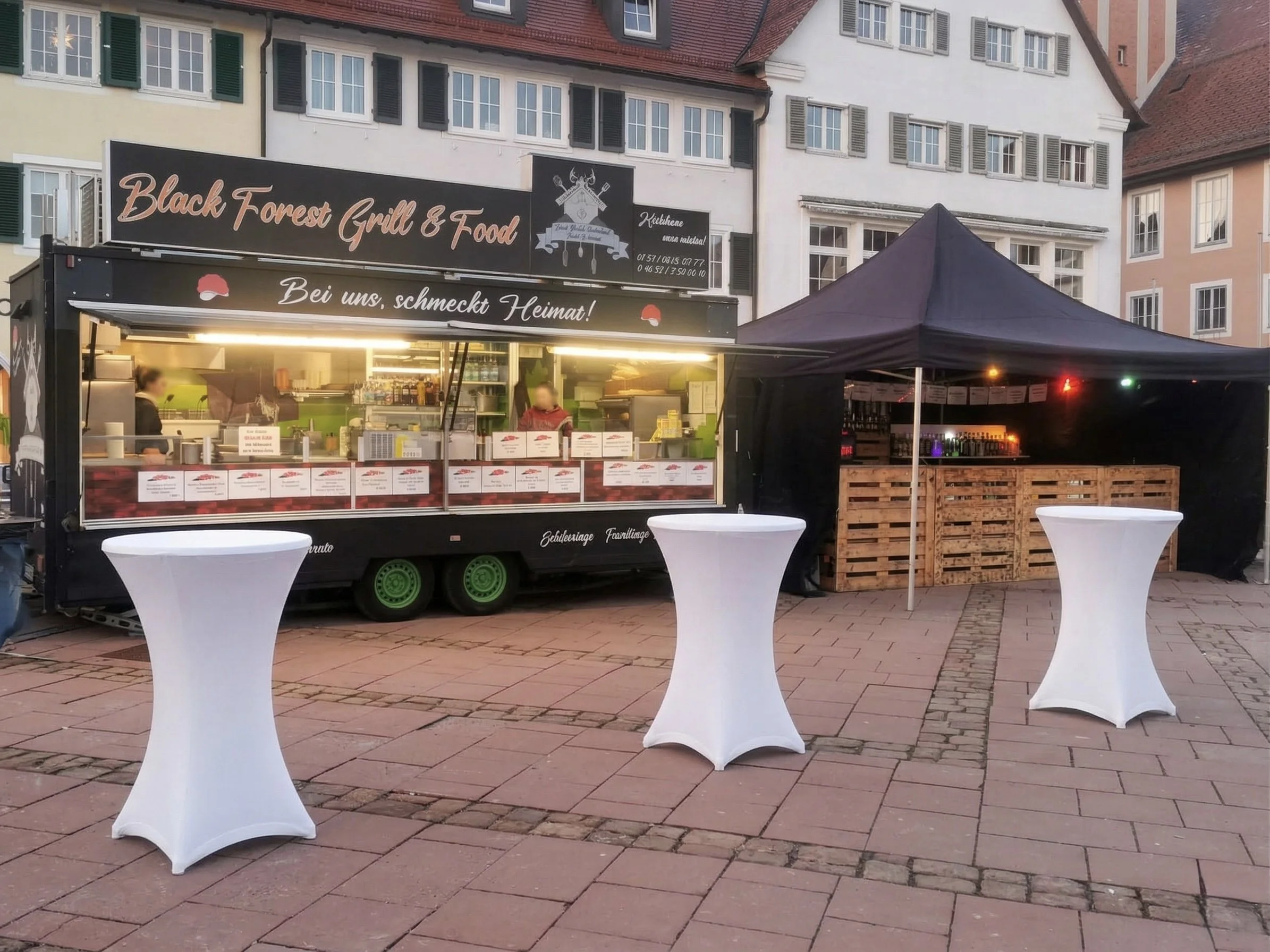 Straßenverkaufsstand für Grill- und Fleischgerichte mit weißen Stehtischen vor einem mehrstöckigen Mehrfamilienhaus. Der Stand trägt den Schriftzug "Black Forest Grill & Food" und den Slogan "Bei uns schmeckt Heimat!"