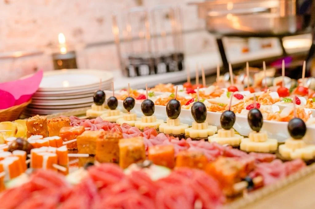 Verschiedene kalte Tapas und Häppchen auf einem Buffet, darunter Käse mit Trauben, Schinken, Sandwiches und andere kleine Snacks.