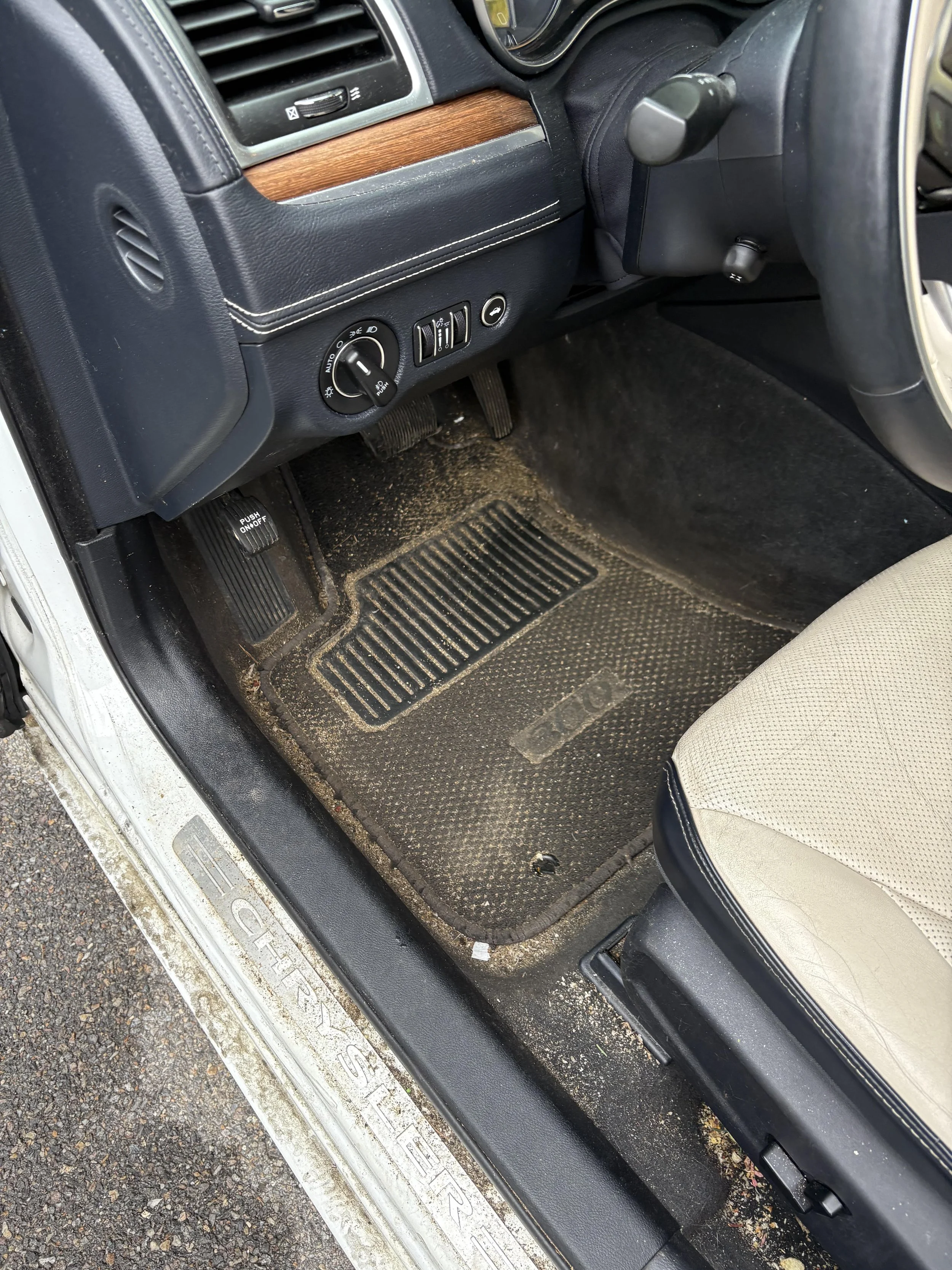 The image shows the driver's side interior of a vehicle, including the footwell with dirty floor mat, accelerator and brake pedals, part of the seat, and the lower dashboard with controls and a wooden trim.