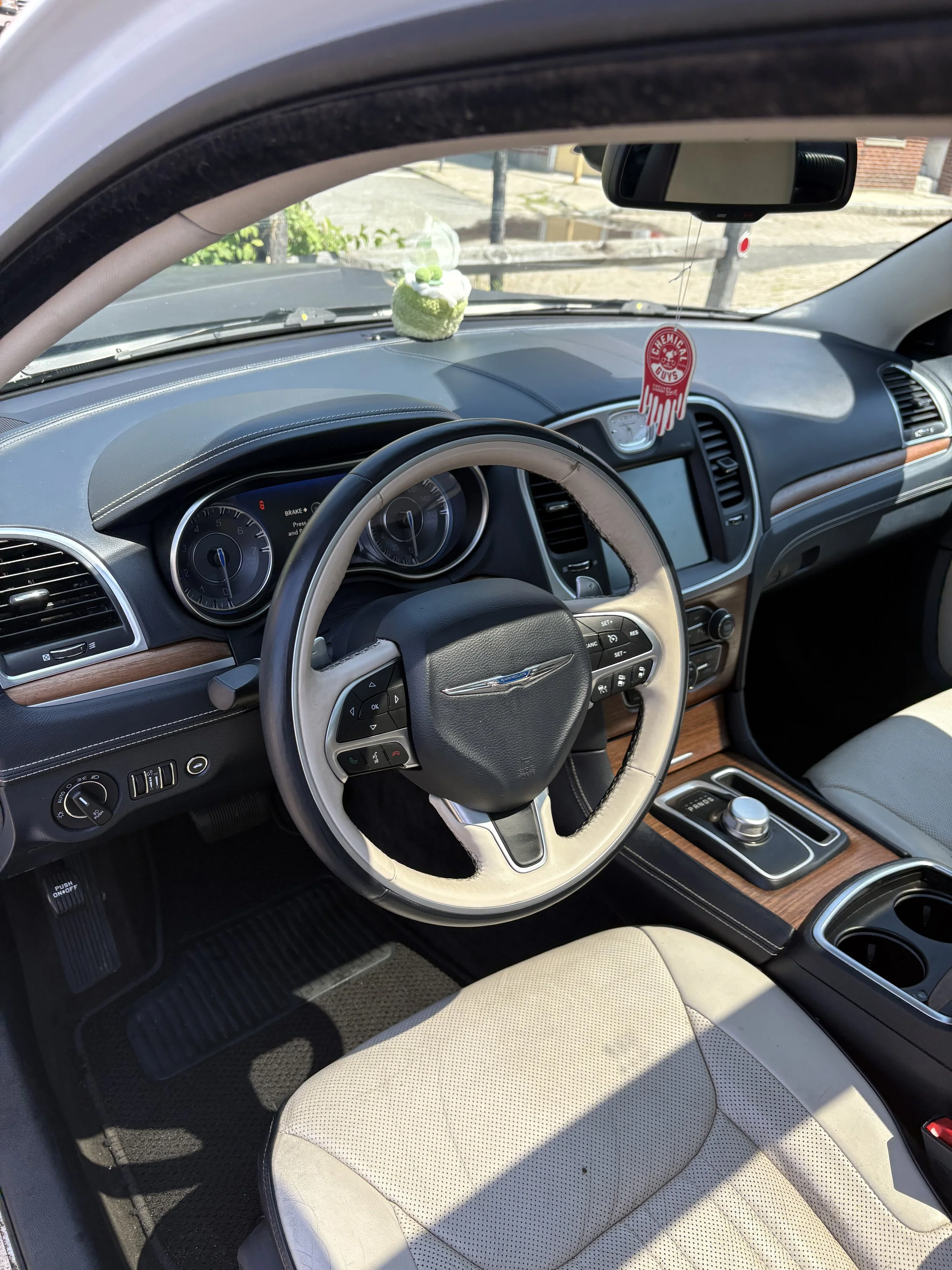 Interior of a car showing the driver's seat, steering wheel, dashboard, and part of the front passenger seat. A small decoration hangs from the rearview mirror, and a plush toy is placed on the dashboard.