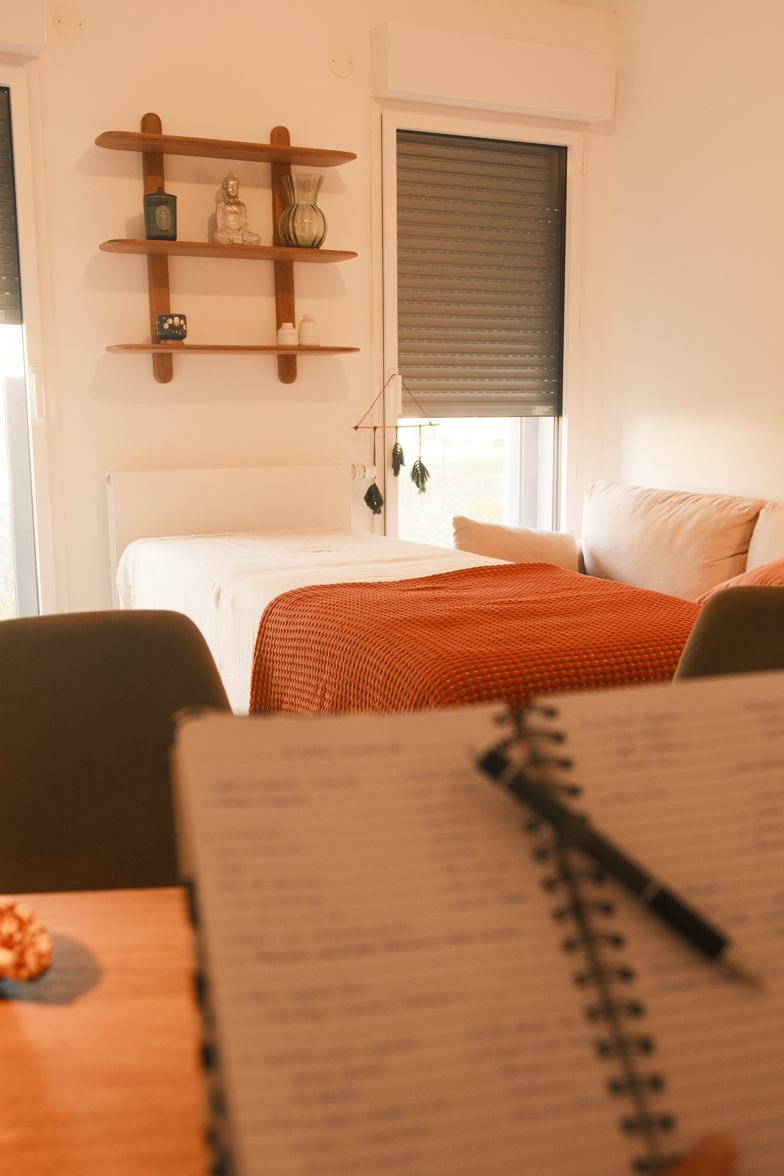 Une chambre avec un lit, une étagère en bois avec des objets décoratifs, une fenêtre avec un volet roulant. Un carnet est visible au premier plan.