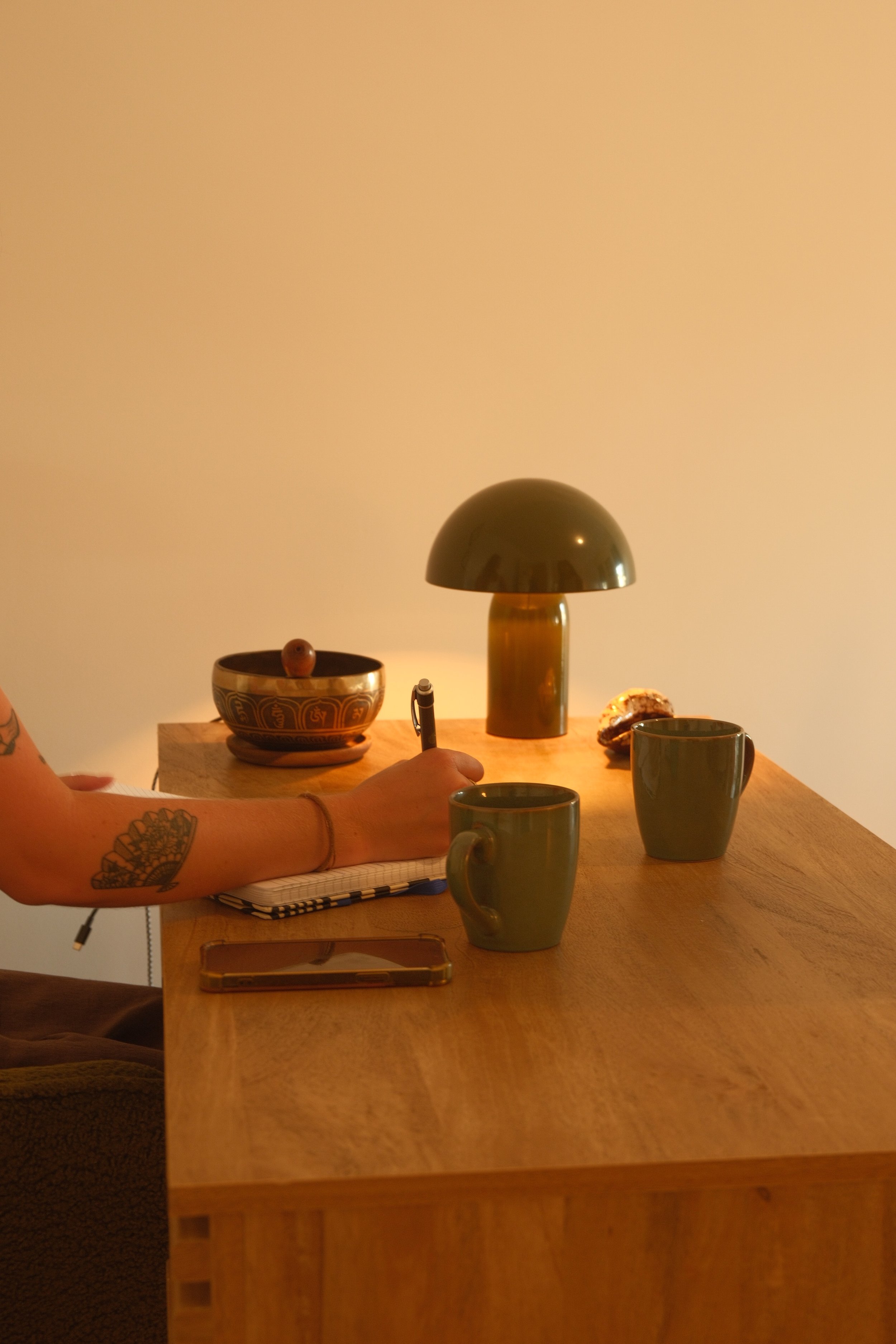 Une table en bois avec deux tasses vertes, un bol décoratif, une lampe de table verte, une personne tenant un stylo et un carnet, autres objets de décoration sur la table.