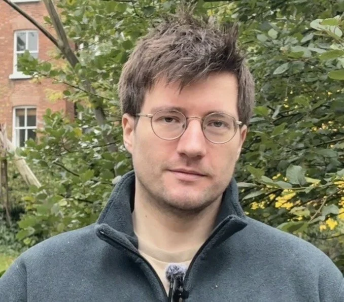 A young man with light skin, short brown hair, and round glasses standing outdoors in front of green foliage and a brick building.