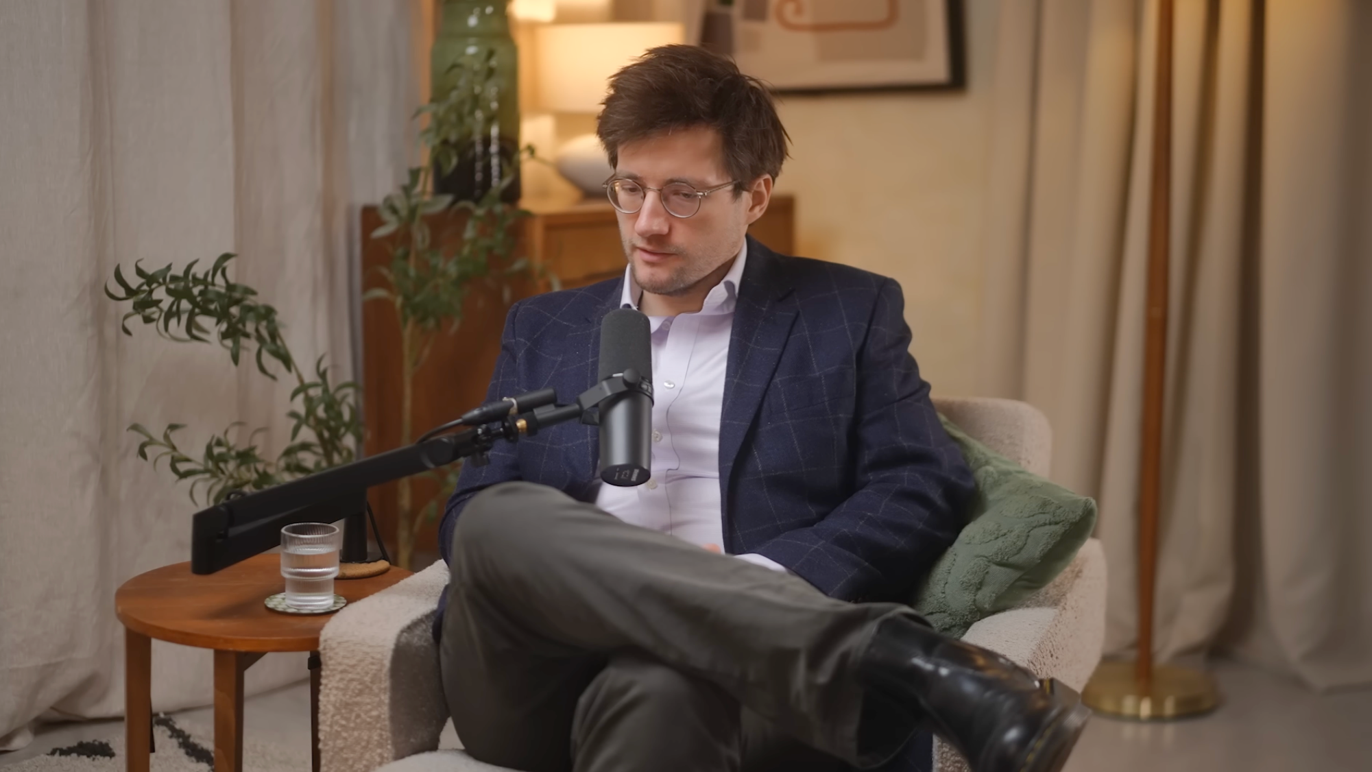 A man with dark hair, glasses, wearing a blue plaid blazer and white shirt, is sitting on a gray sofa, speaking into a microphone. He has one leg crossed over the other and is looking down. There is a small round wooden table in front of him with a glass of water. The background features a cozy living room with a lamp, green plant, and framed artwork on the wall.
