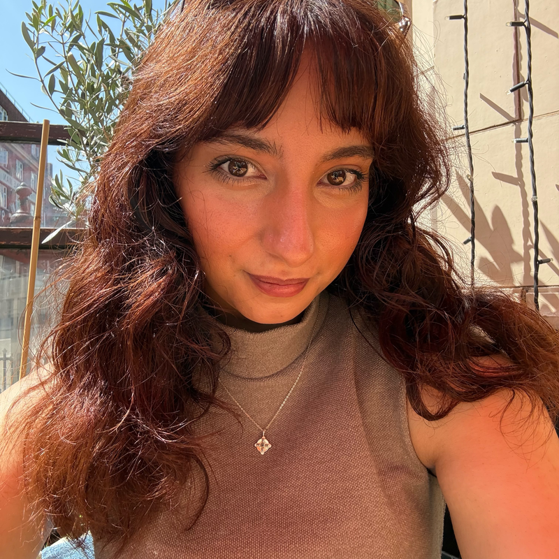 A woman with long, curly brown hair and brown eyes, wearing a sleeveless top and a necklace with a small pendant, standing outdoors on a sunny day.