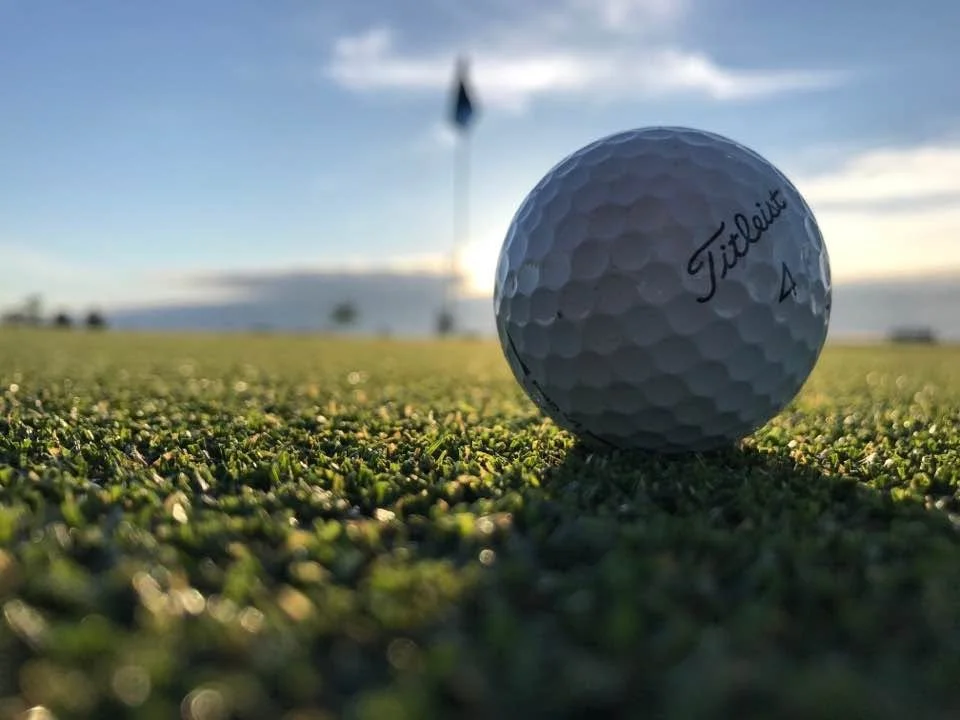 Close-up of a golf ball on a putting green with a flag in the background during sunset.