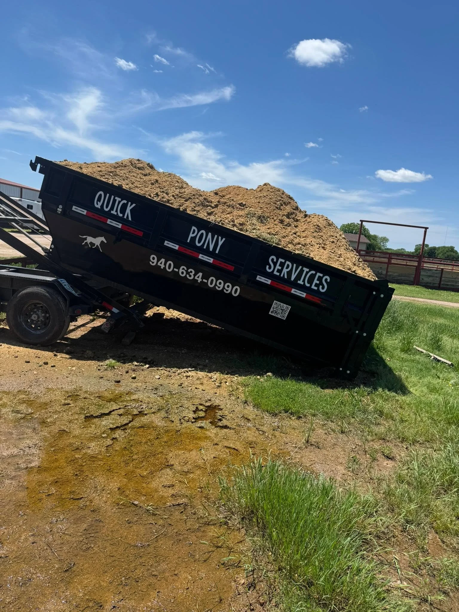 A black utility trailer filled with dirt and soil, with the words 'QUICK PONY SERVICES' and a contact number on its side, parked on a grassy area under a blue sky with scattered clouds.