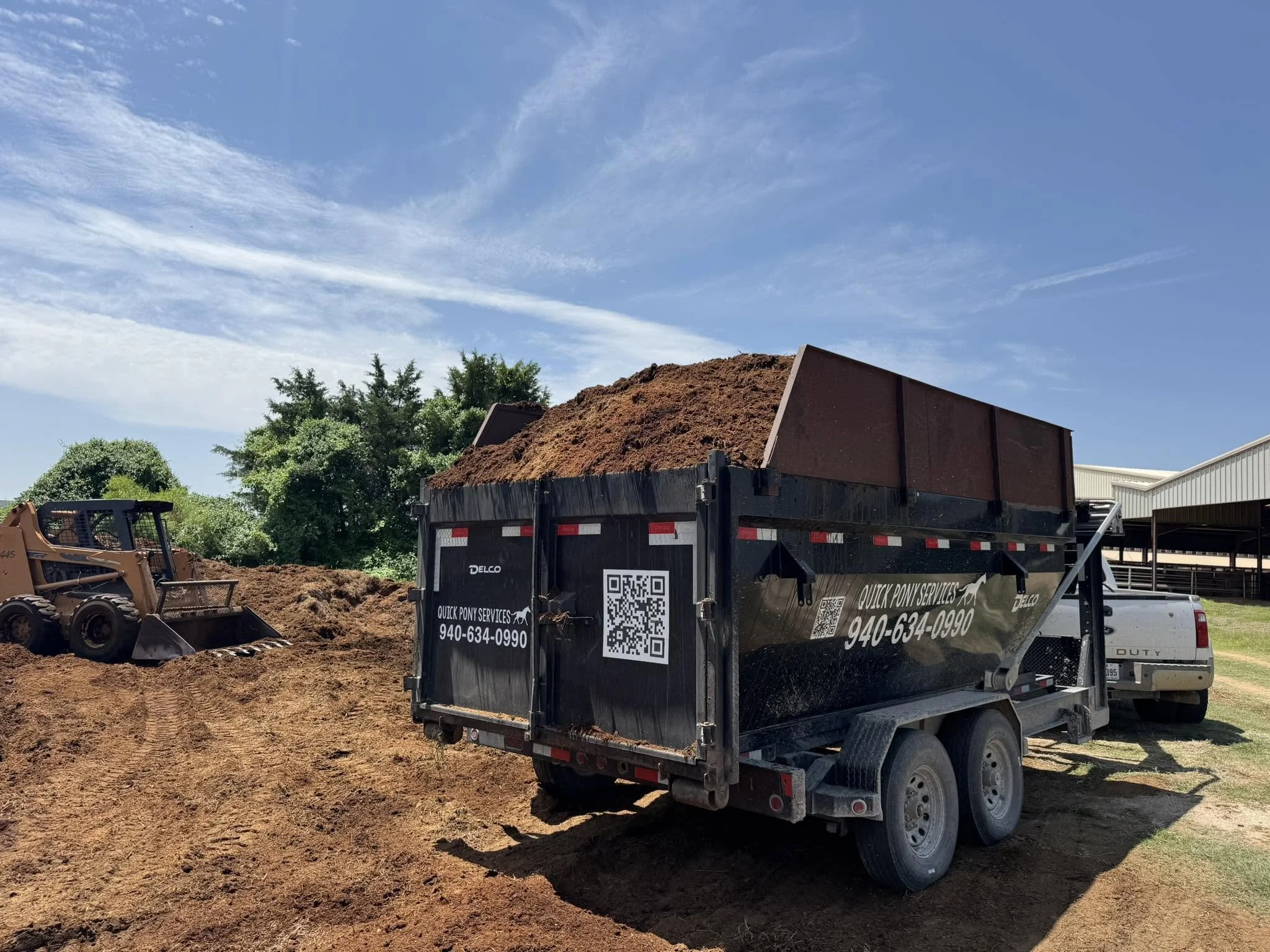A construction site with a black dump truck filled with reddish dirt, a small bulldozer, trees, and a building in the background under a blue sky with wispy clouds.