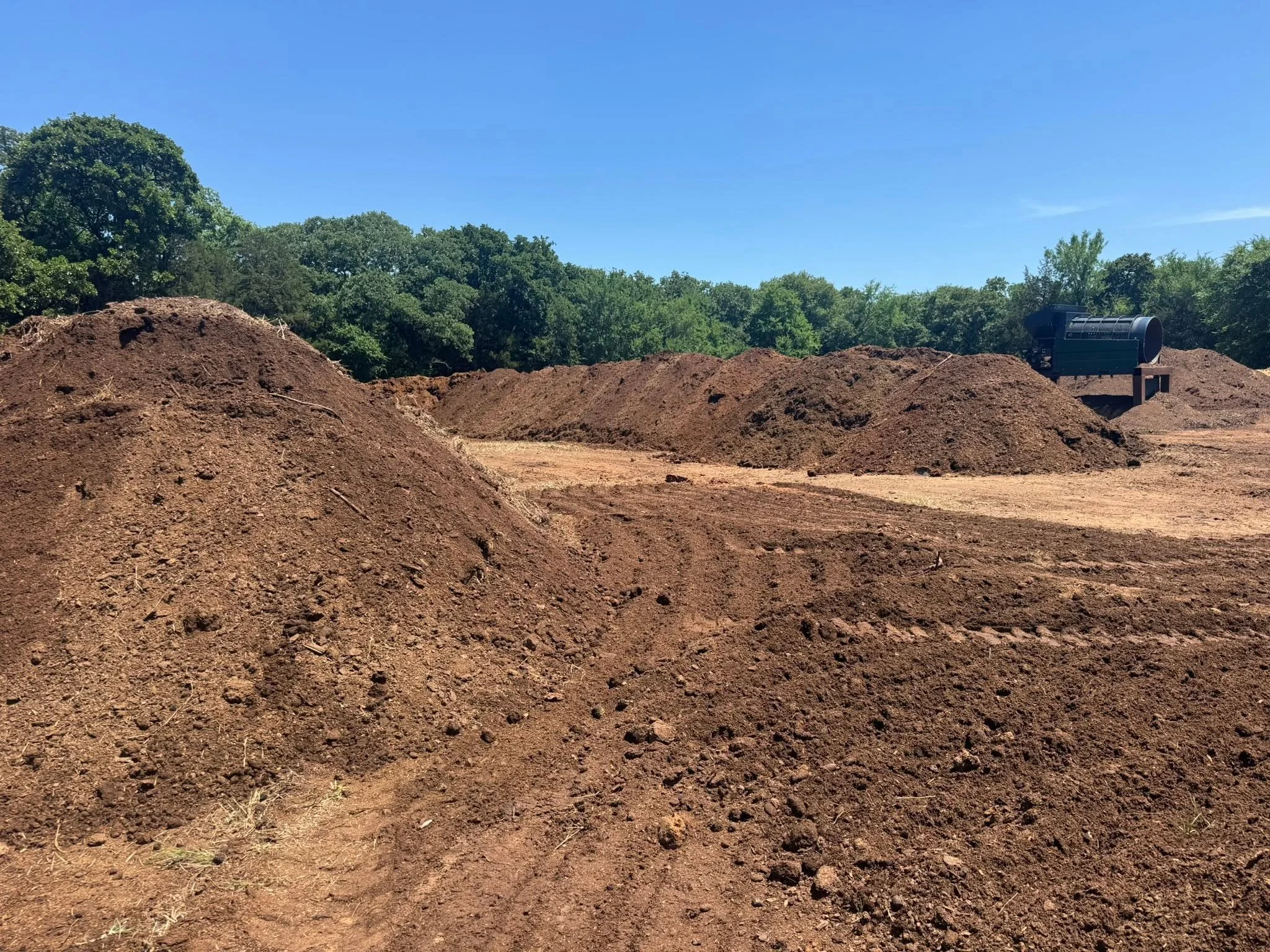 A construction site with large piles of dark brown soil and dirt, a piece of industrial equipment or machinery, and a backdrop of green trees under a clear blue sky.