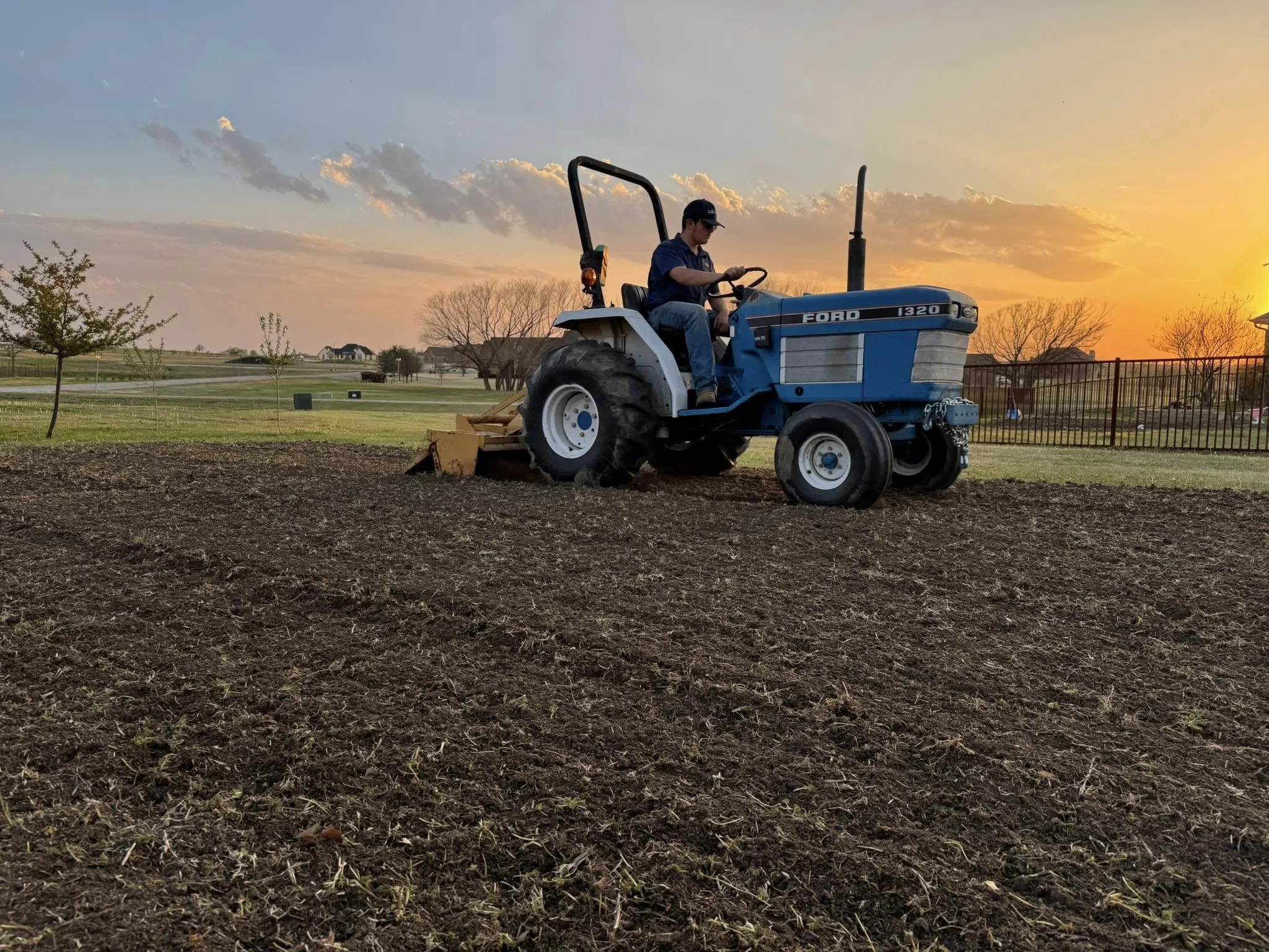 A man operating a blue Ford tractor on a farm during sunset, tilling a garden with trees and a fence in the background.