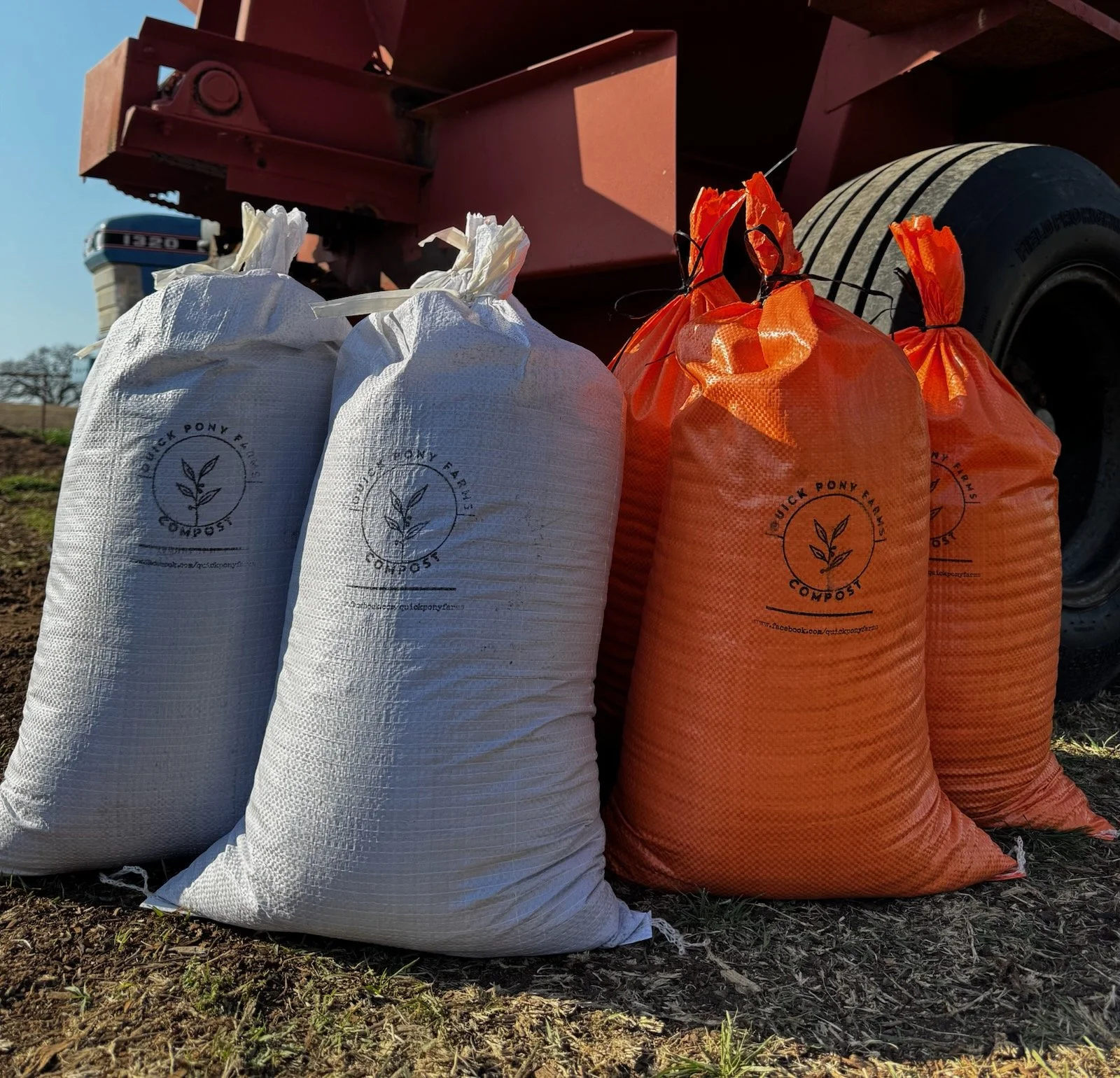 White and orange compost bags stacked outdoors near agricultural machinery on dirt ground
