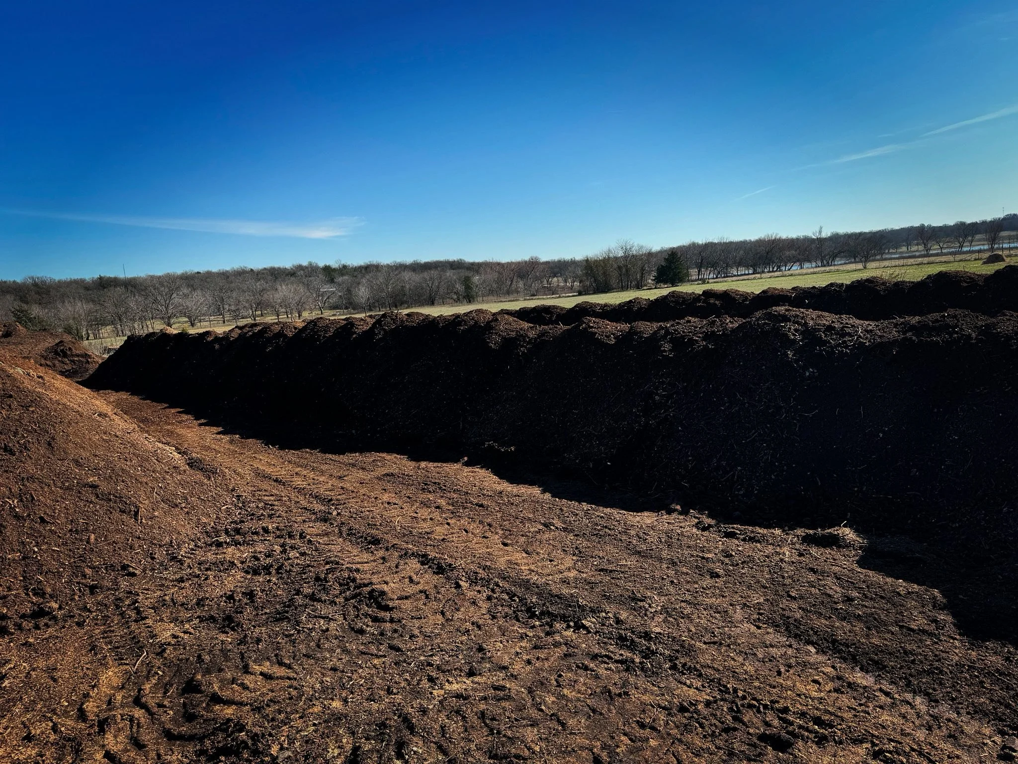 A landscape showing freshly turned dark brown soil in the foreground, a grassy field with leafless trees in the middle ground, and a clear blue sky in the background.