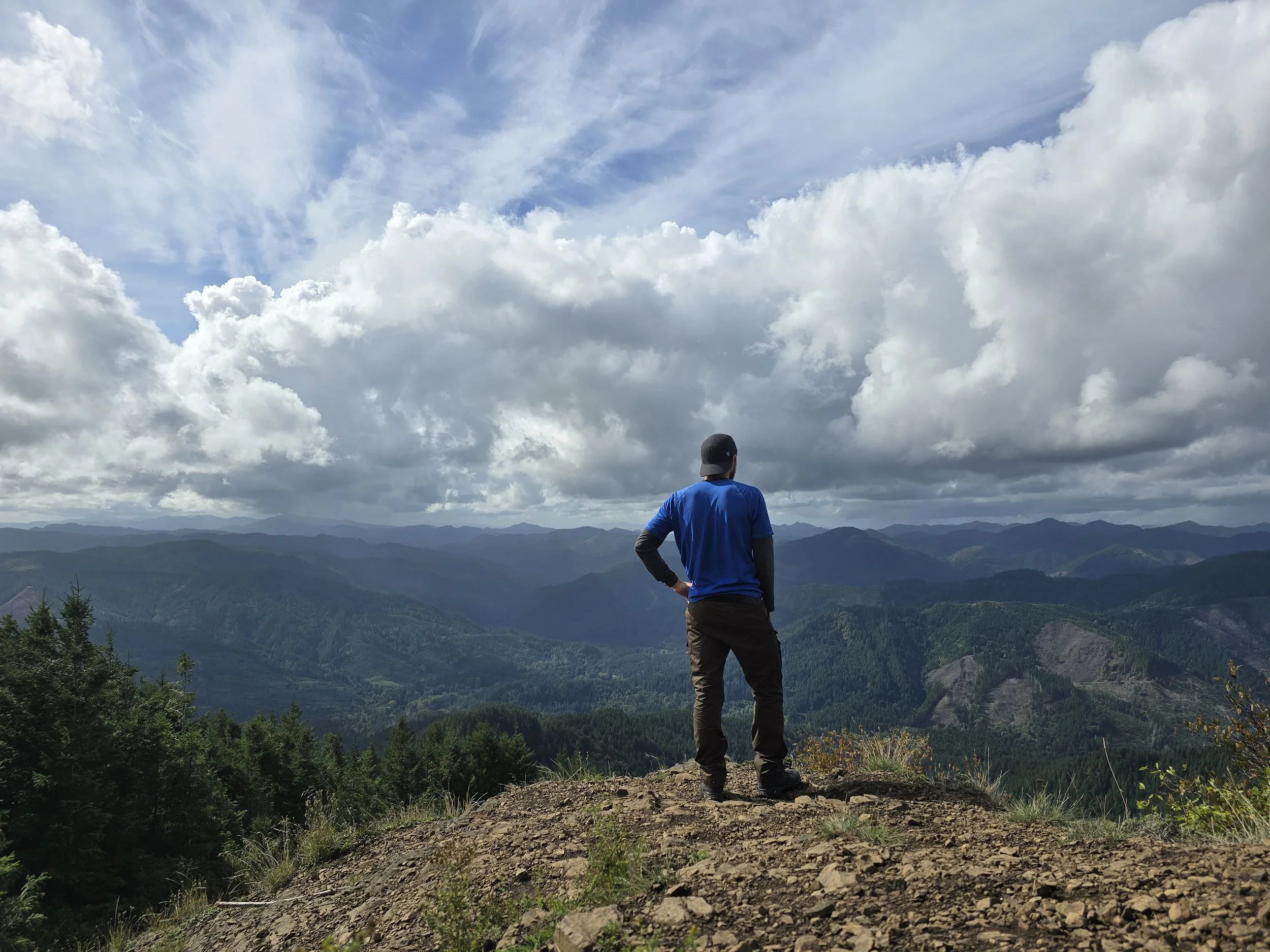A person standing on a mountain overlooking a scenic view of rolling hills and valleys under a partly cloudy sky.