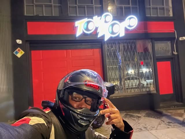 A person taking a selfie in front of a store called 'Togo' at night. The person is wearing a motorcycle helmet and sunglasses, and is making a peace sign. The store has a red door and window frames with a barred window, and the store sign is brightly lit.