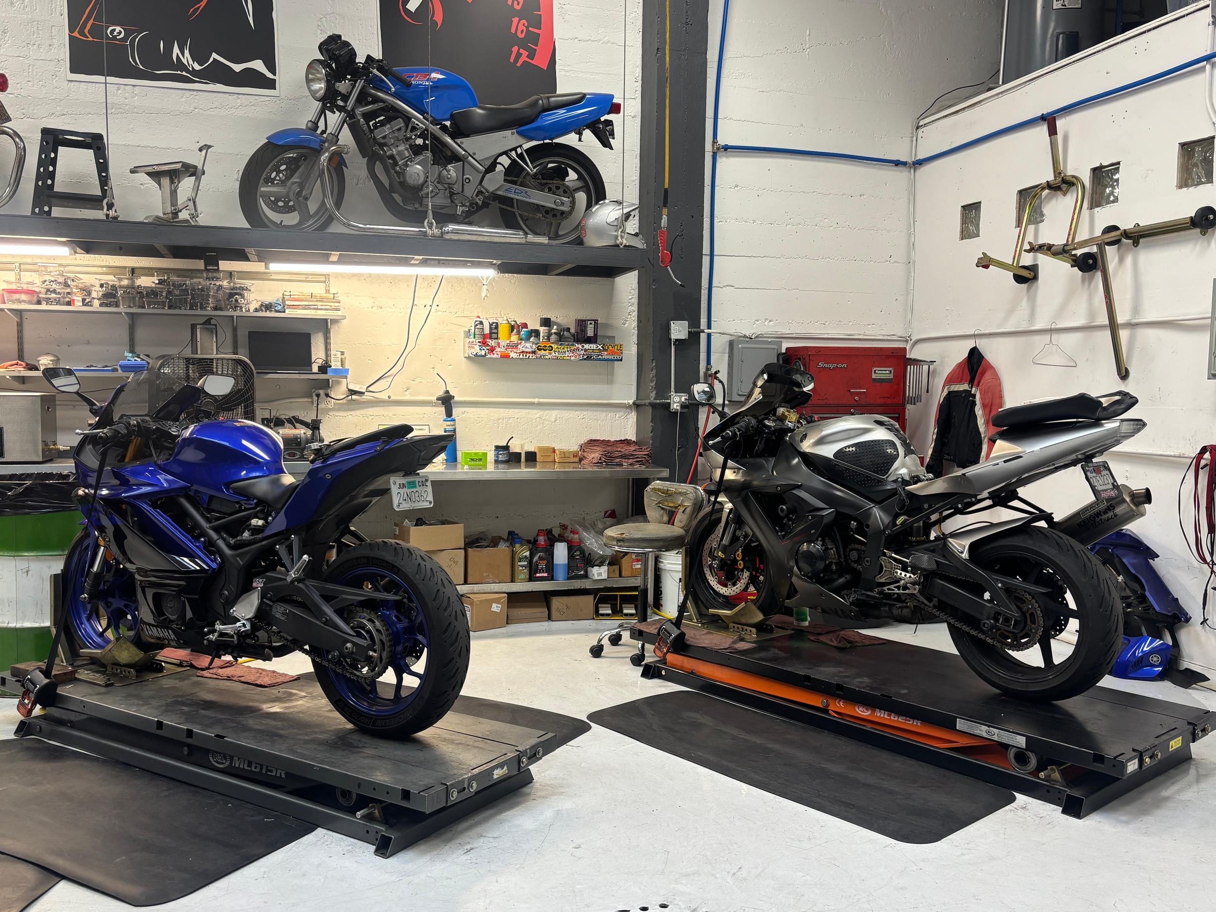 A garage with two motorcycles, one on a lift and the other on a stand, surrounded by tools, shelves with supplies, and various equipment.