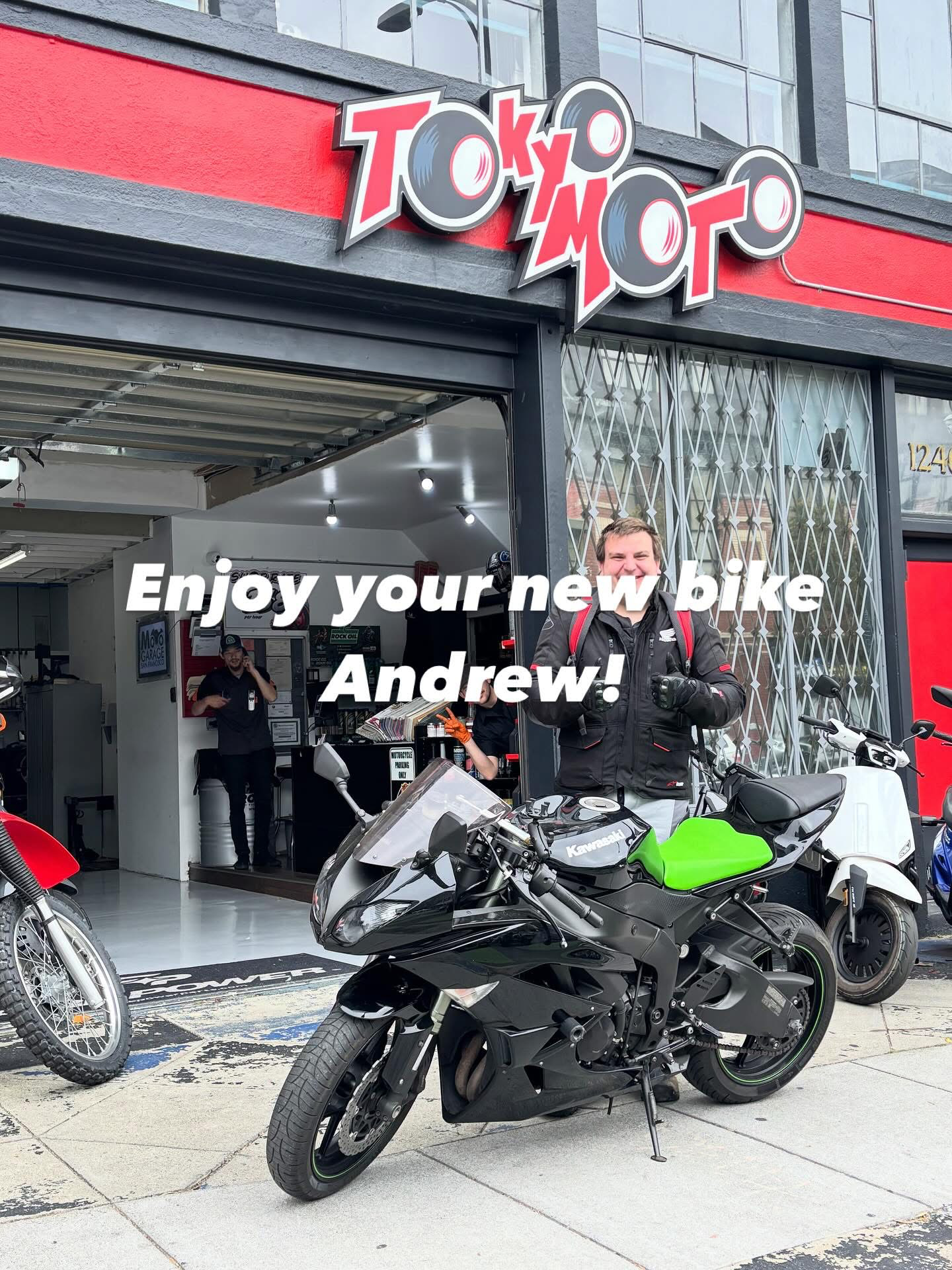 A motorcycle dealership called Tokyo Moto with a sign above the entrance. A man in motorcycle gear stands next to a black motorcycle with green accents. Inside, a staff member is visible behind the counter. Several motorcycles are parked outside on the sidewalk.