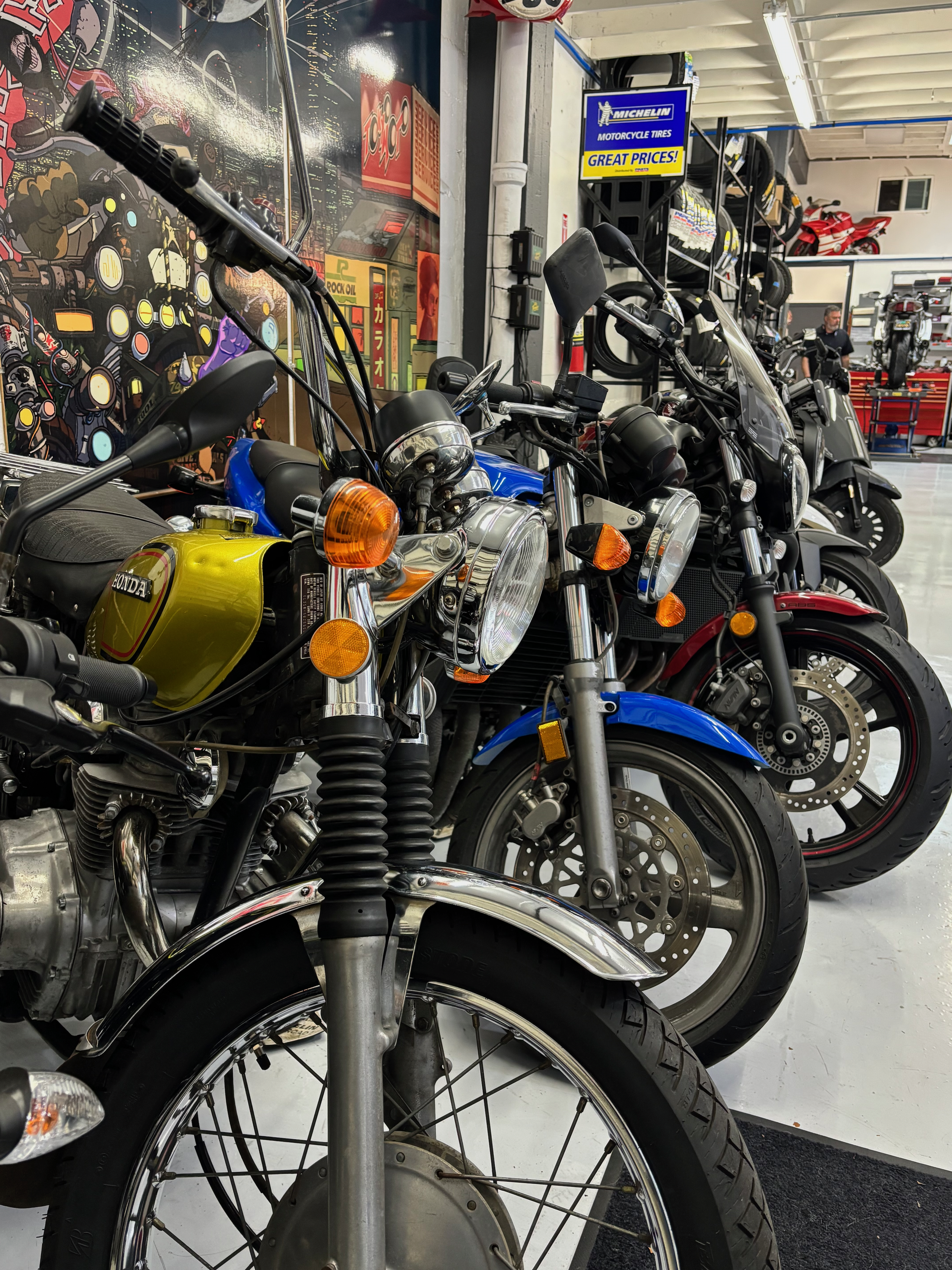 A row of motorcycles parked inside a showroom with various bike models and a colorful background poster.