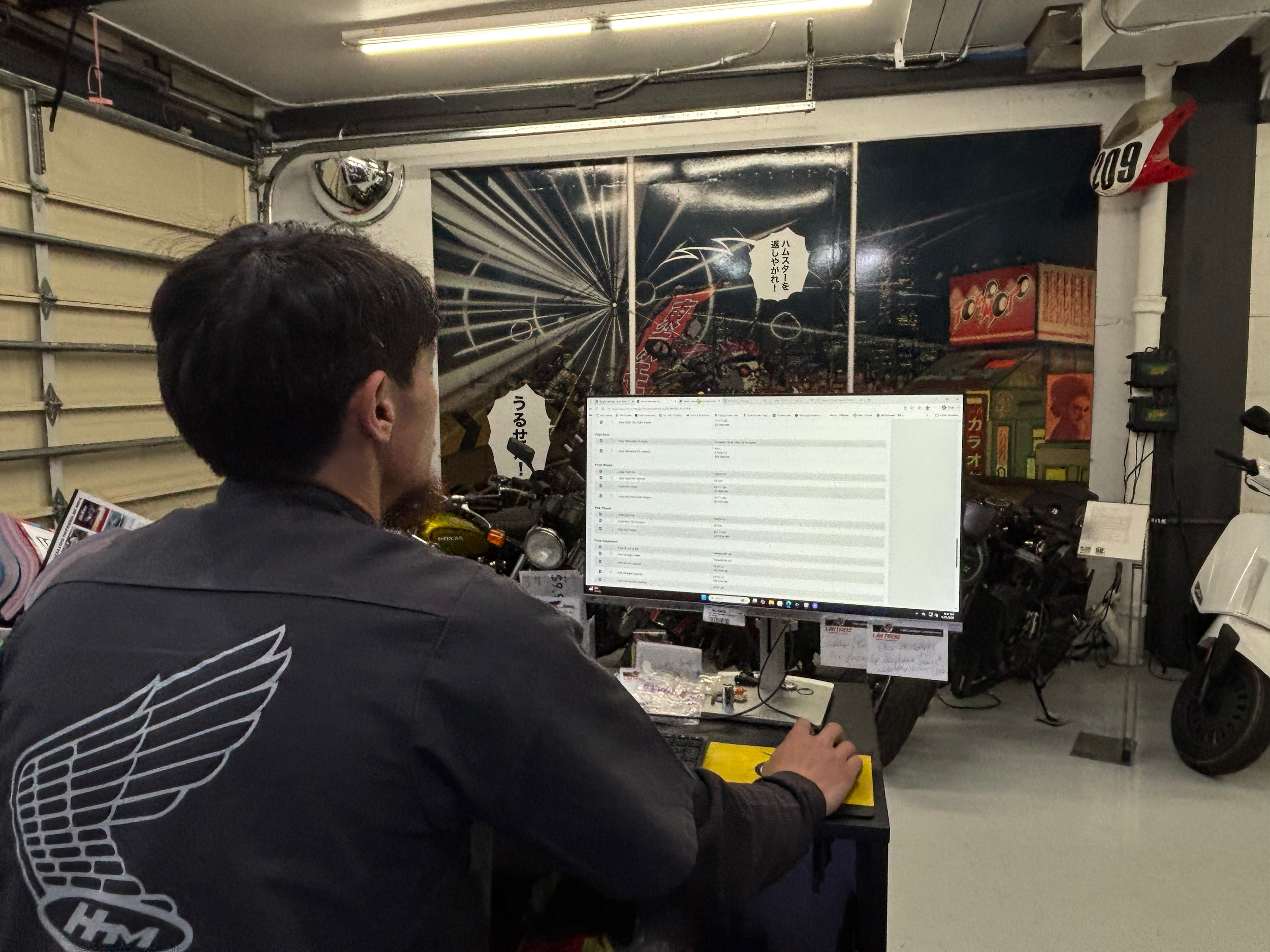 Man sitting at a desk with a computer monitor, in a garage or workshop. Behind him, there is a motorcycle and some posters or artwork on the wall, including a dartboard and Japanese text.