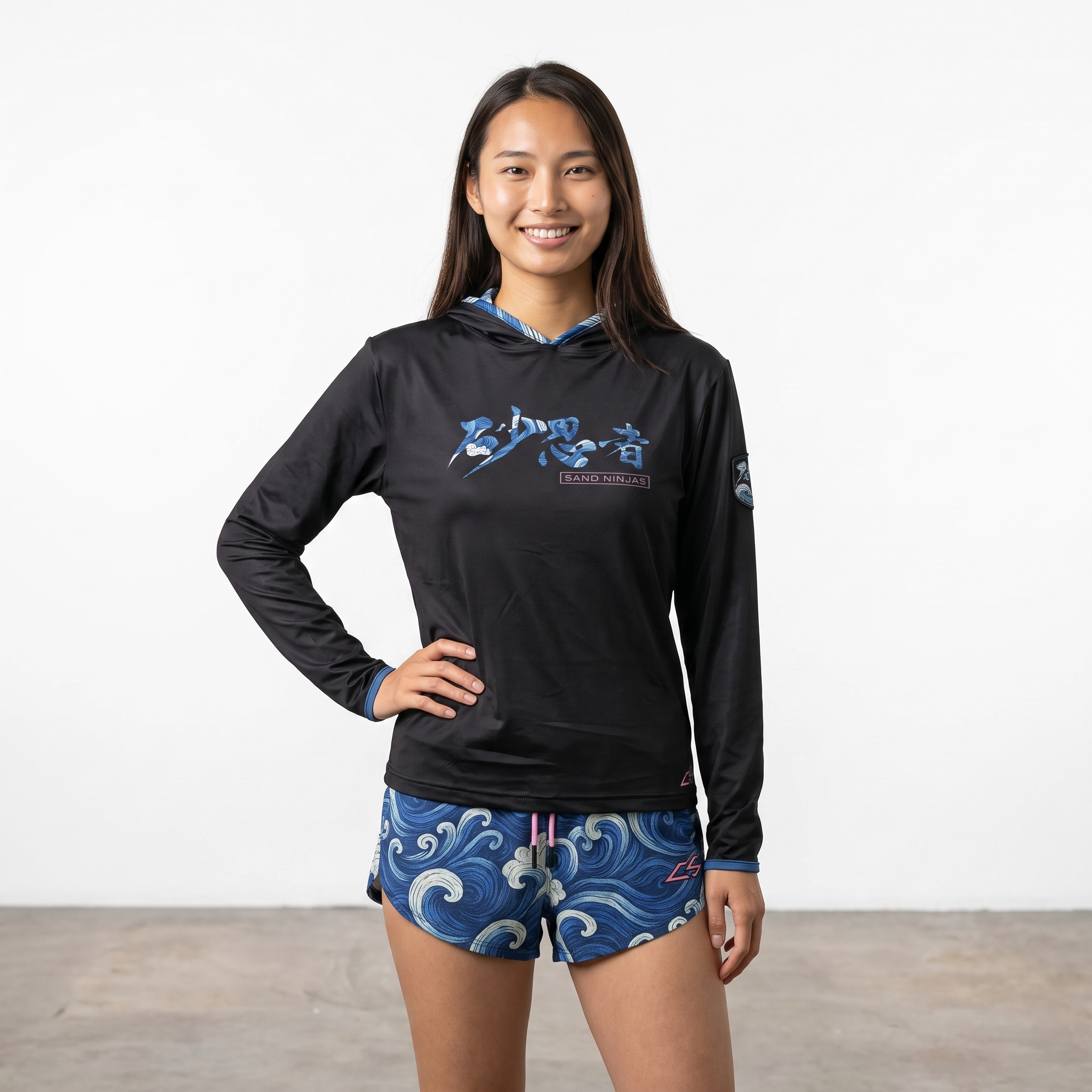 Young woman wearing a black long-sleeve sports shirt with Japanese characters and "Sand Ninjas" logo, and blue wave-patterned shorts, stands with her hand on her hip against a plain white background.