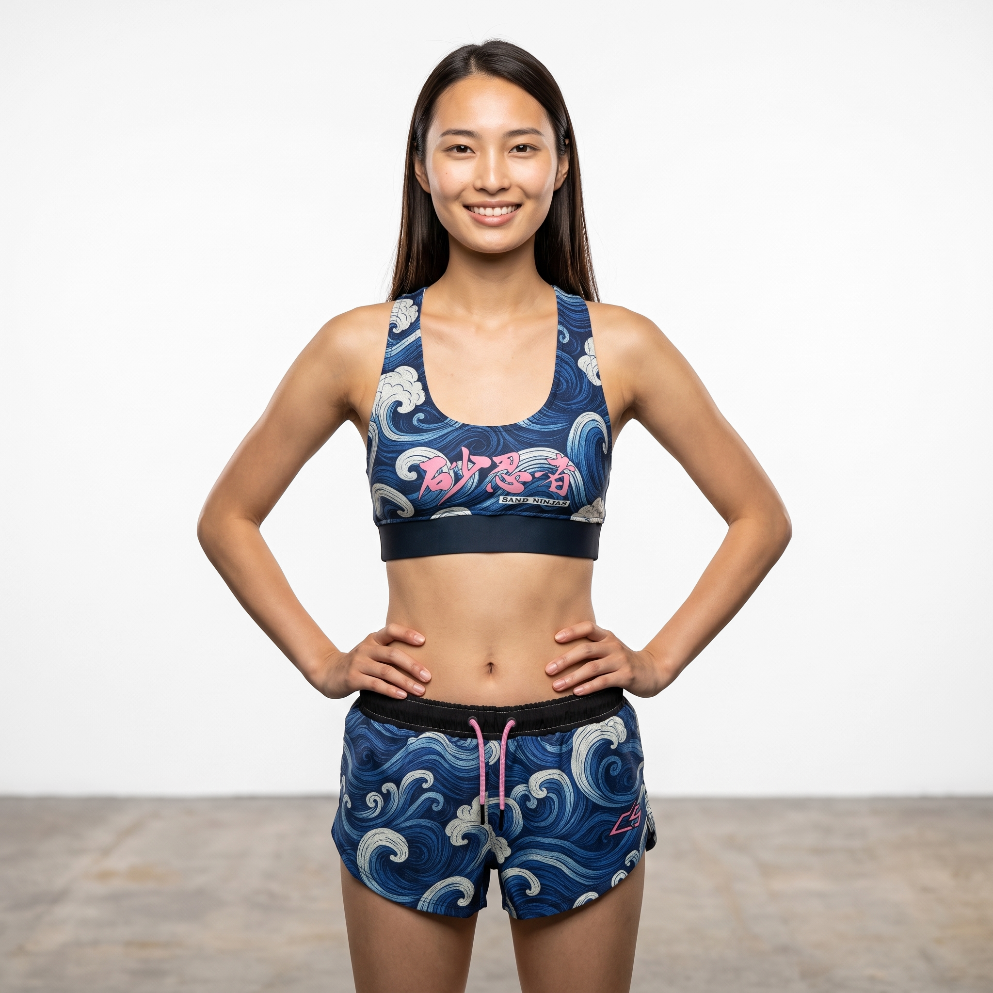 A young woman in athletic clothes stands with her hands on her hips in a gym or studio, smiling at the camera.