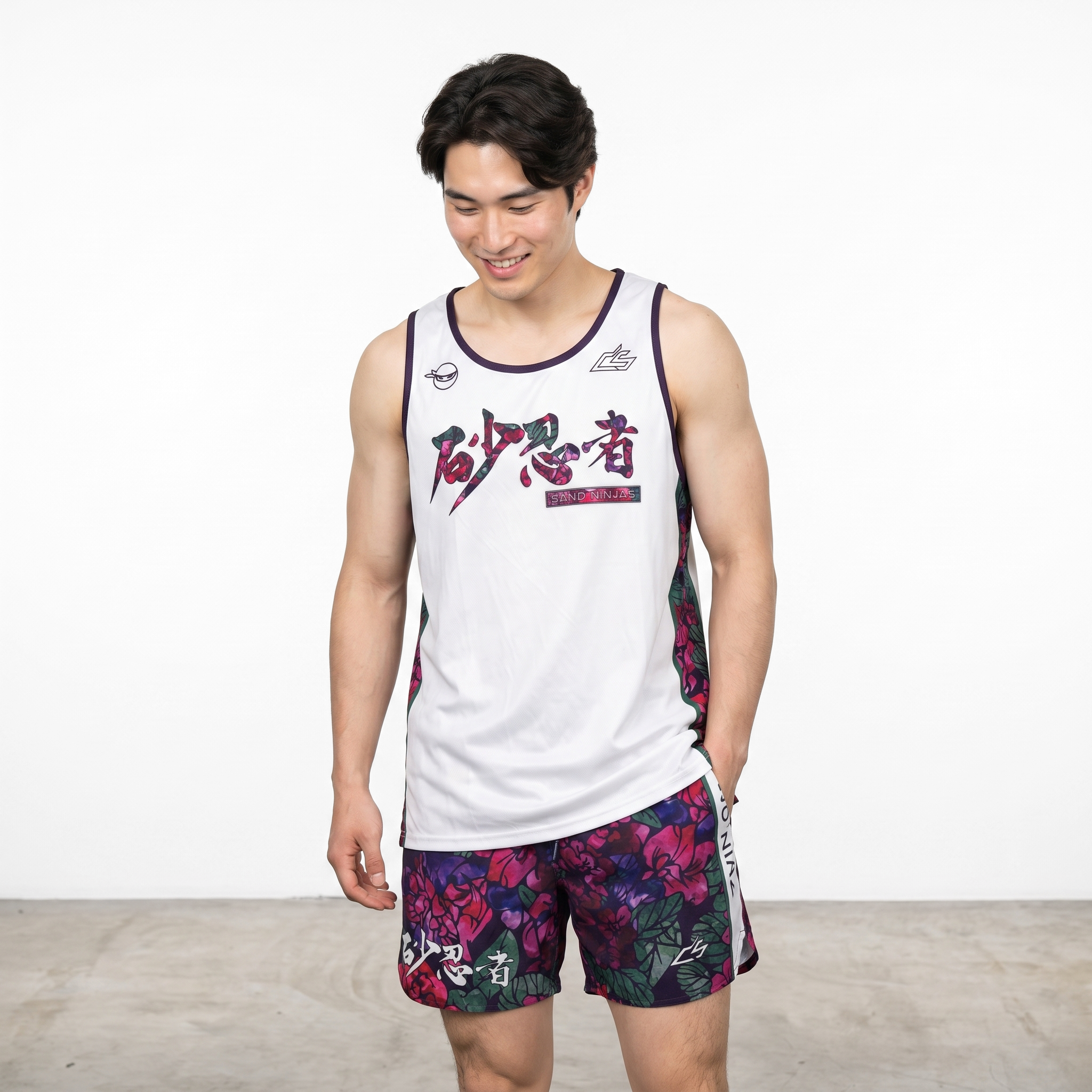 Young man in a sleeveless athletic shirt and colorful patterned shorts smiling and looking down, standing against a plain white background.