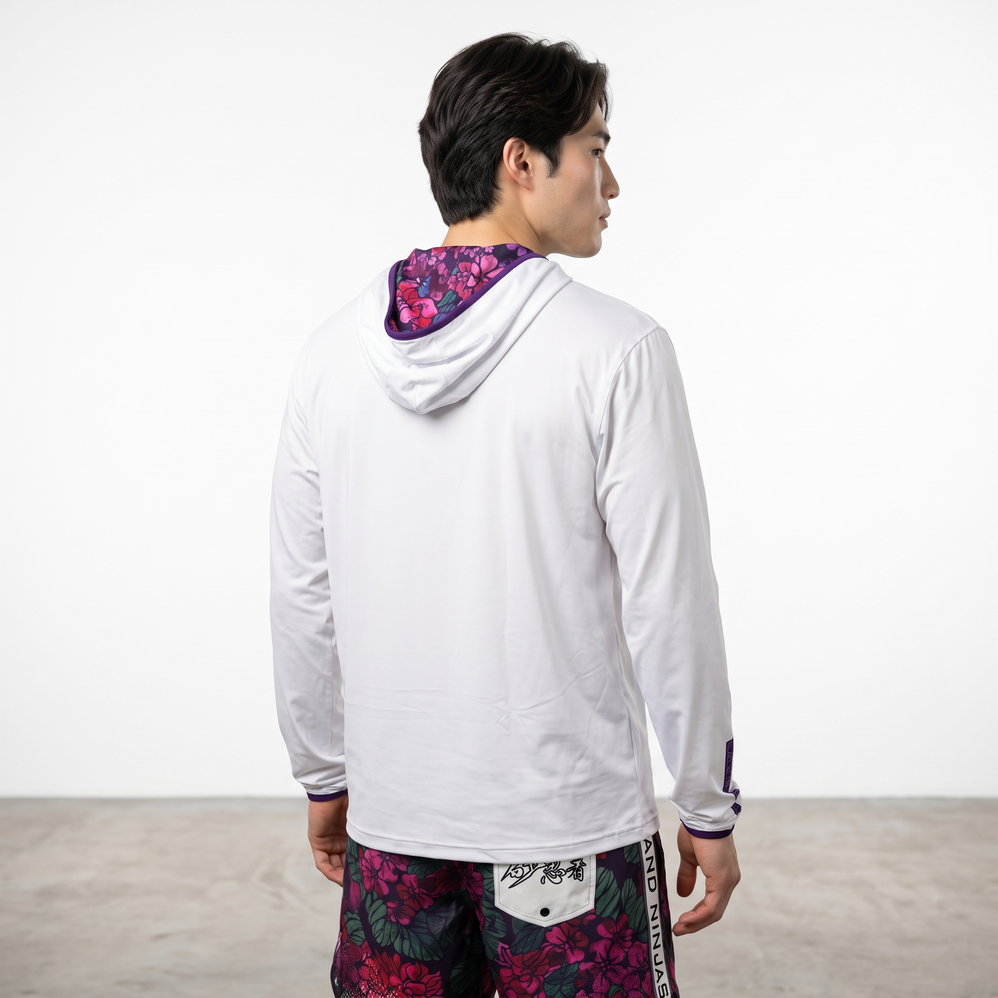 Side profile of a young man with dark hair wearing a white jacket with purple accents and floral-patterned shorts, standing against a plain white background.