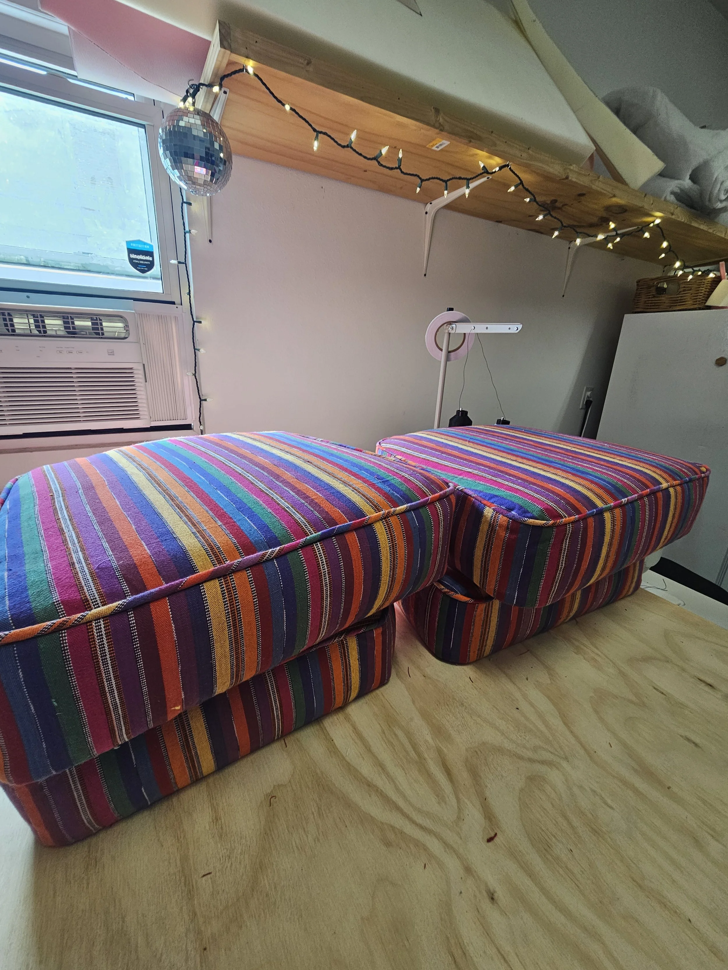A colorful striped couch is in the foreground, with a wooden table above it supported by white brackets. Fairy lights are hanging from the table, and a disco ball is nearby. There's a window with a window air conditioning unit beneath it on the left 